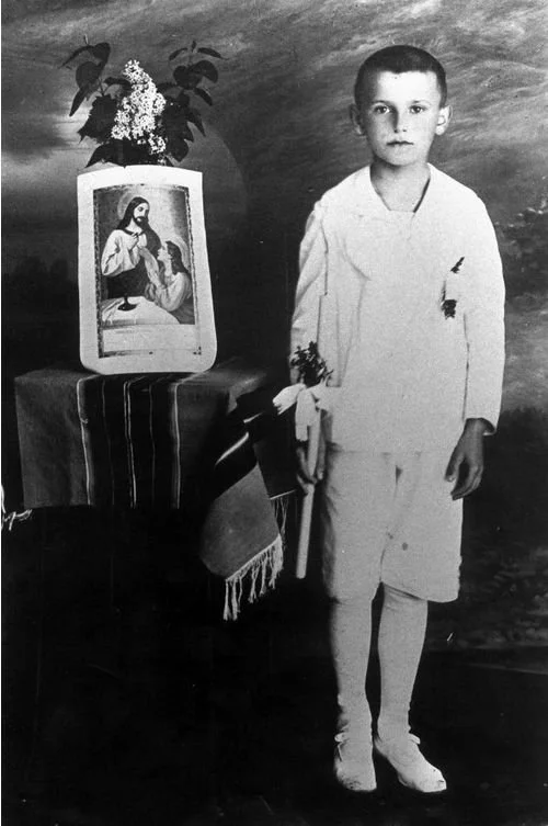 Black and white photo of a young boy standing next to a table with a religious picture and a plant, holding a flower in his hand, wearing a white outfit.