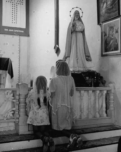 A woman and a young girl kneel and pray at a Catholic altar featuring a statue of the Virgin Mary. The room is decorated with religious posters and framed images.