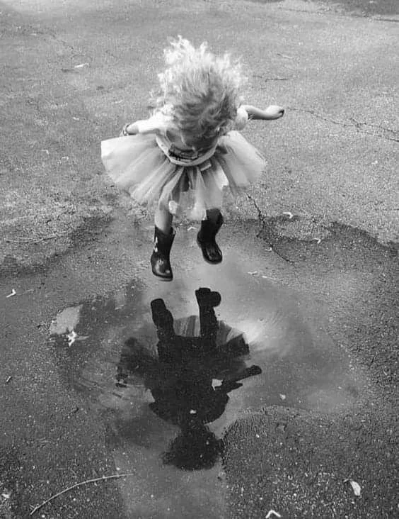 A young girl in a tutu and rain boots is jumping over a puddle, creating a reflection of herself in the water.