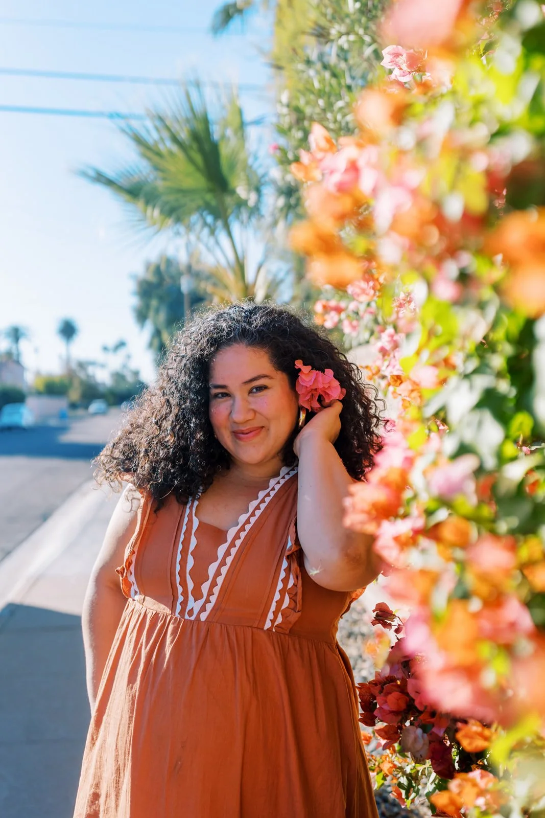 A woman with curly dark hair holding a pink flower near her face, standing next to a wall of colorful pink and orange flowers, with palm trees and a clear blue sky in the background.