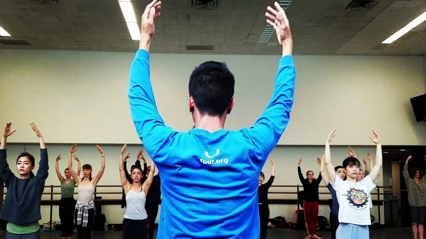 A dance instructor leading a dance class with students raising their arms in a pose, in a dance studio with mirrors and a ballet barre.