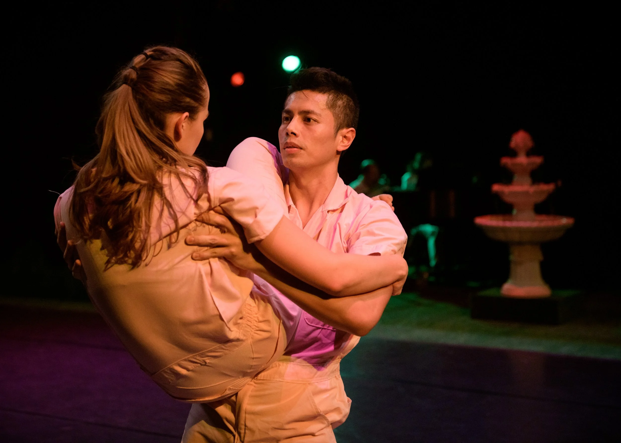 A man is holding a woman in his arms during a dance performance on stage, with a fountain in the background, lit with pink and green lighting.