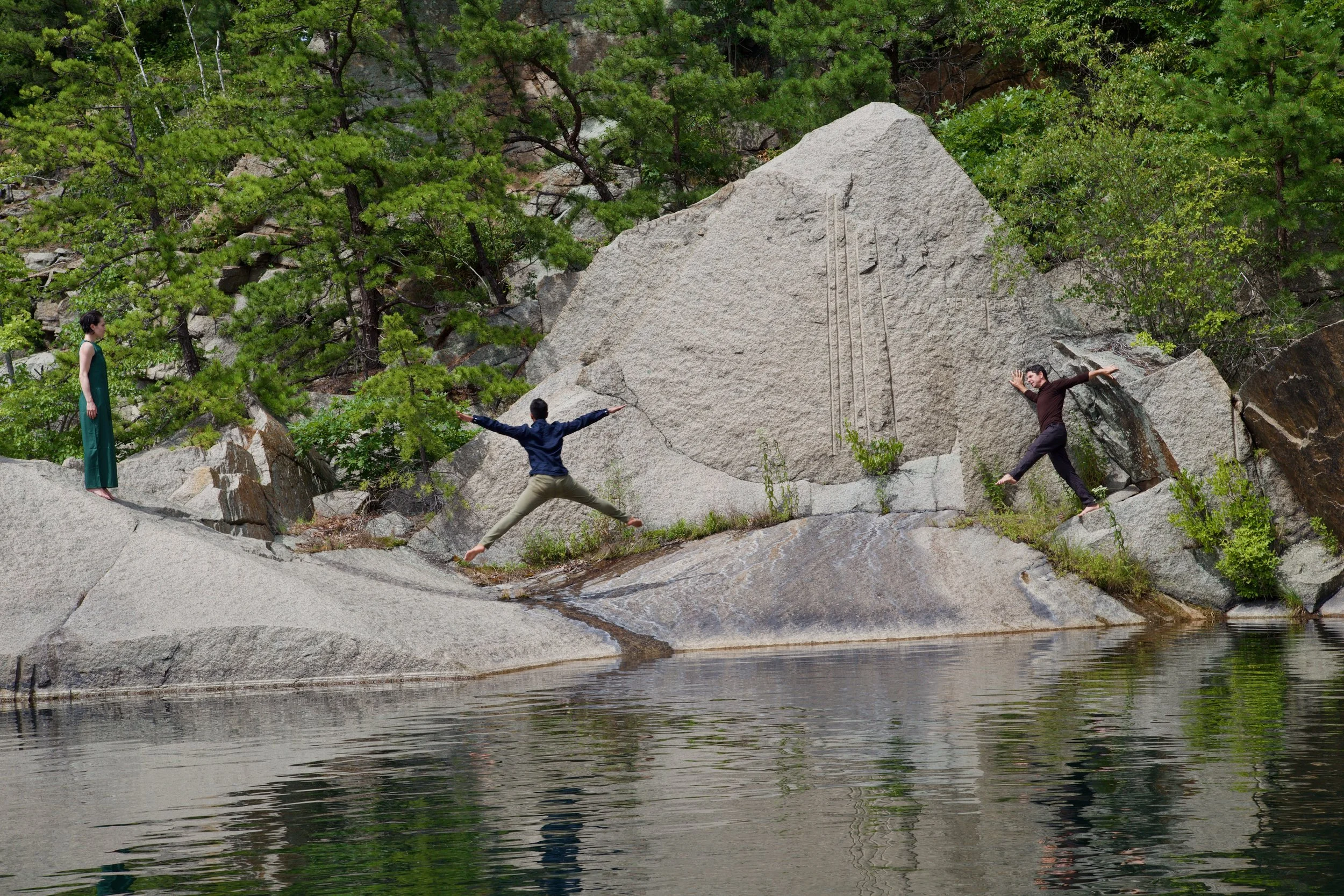 Three people are on large rocks near a body of water. One person in a green dress stands on a rock to the left, observing. Two people are in mid-movement, one jumping between rocks and another crouching behind a large boulder on the right. The background features green trees and bushes.