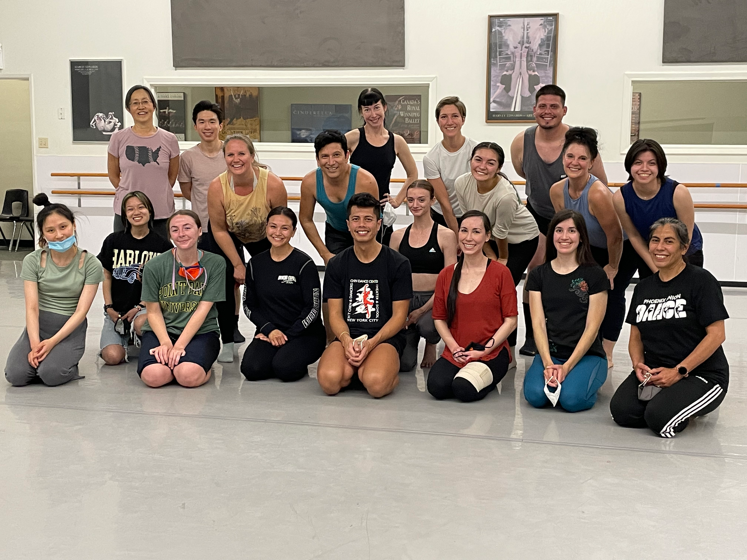 A group of diverse people, mostly young adults, poses in a dance studio after a class. They are smiling, some kneeling and others standing, with ballet barre and posters in the background.