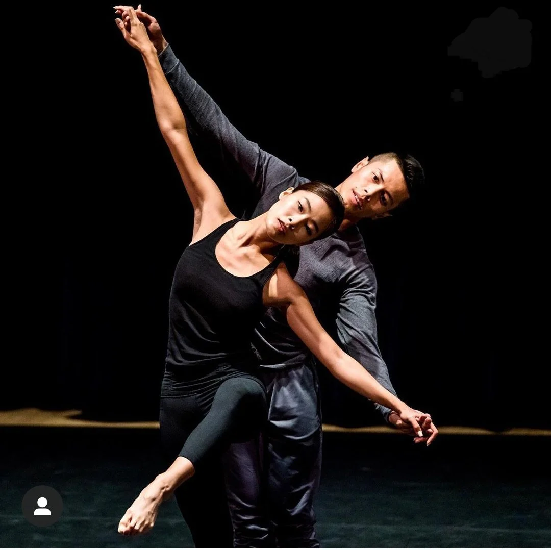 Two dancers, a woman and a man, performing a contemporary dance on a dark stage. The woman is in front, balancing on one foot with her other leg raised and extended, and her head tilted to the side with her eyes closed. The man stands behind her, supporting her with his arm around her waist, and their bodies are intertwined in an expressive pose.