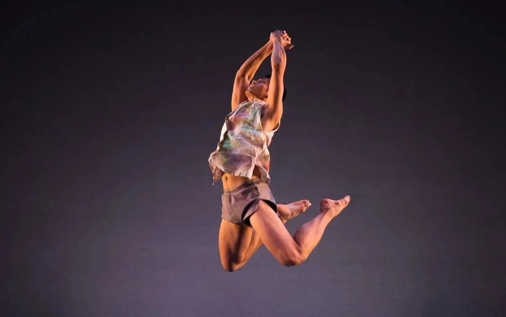 Gary Champi leaps in mid-air against a dark background, with his arms extended above his head and legs bent behind him.