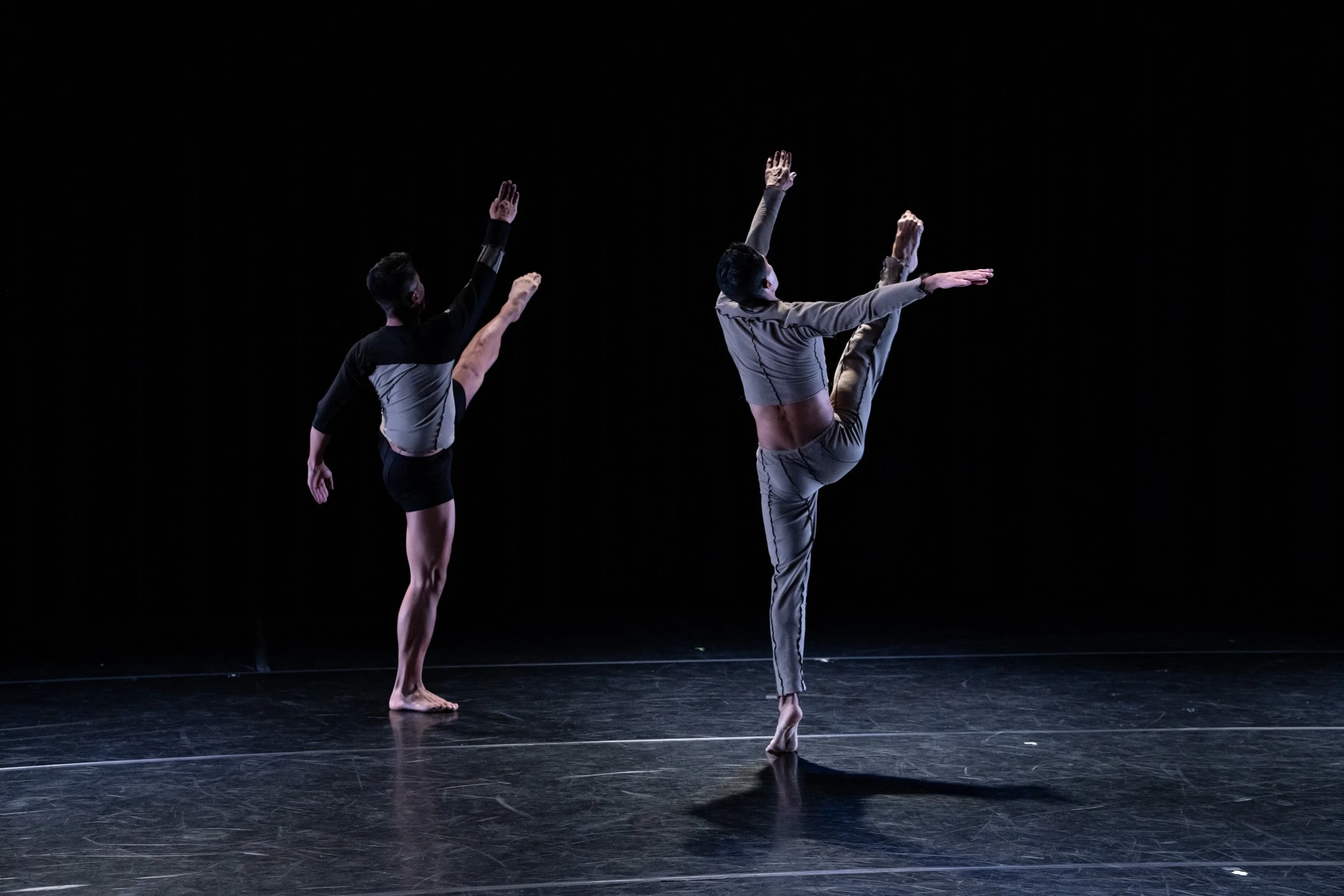 Two dancers are performing on a dark stage with a black background, one balancing on one foot with arms outstretched, and the other with one arm raised and the other extended to the side.