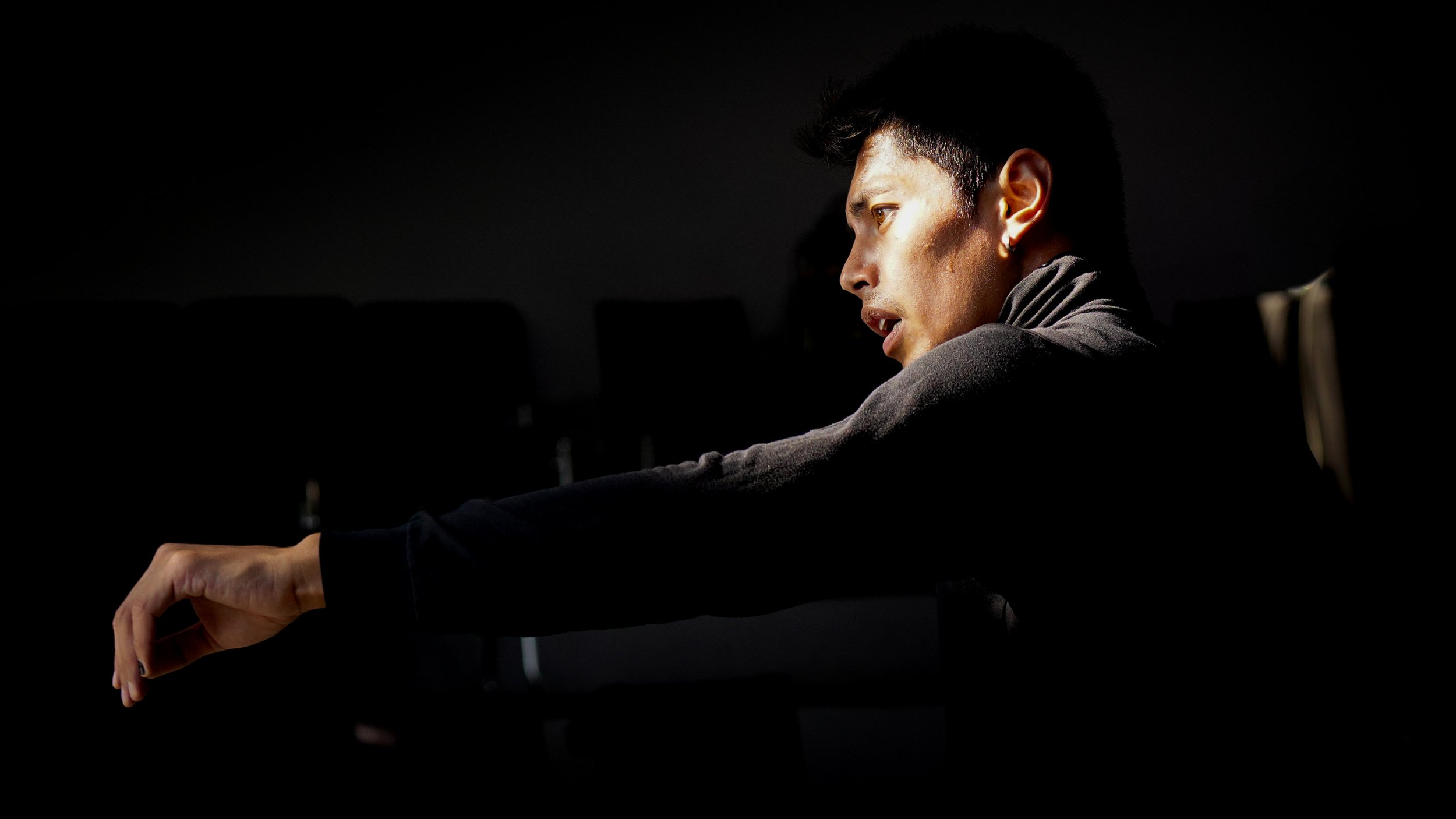 A person with short dark hair sitting in a dark room, reaching forward with their left arm, with a focused expression.