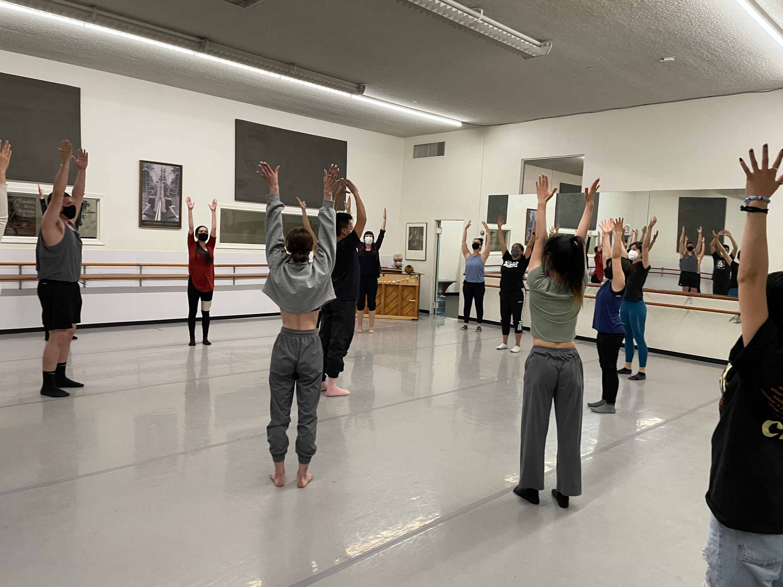 Group of people in a dance studio practicing dance moves with their arms raised, wearing casual and athletic clothing, some with face masks, mirrored wall reflecting their positions.
