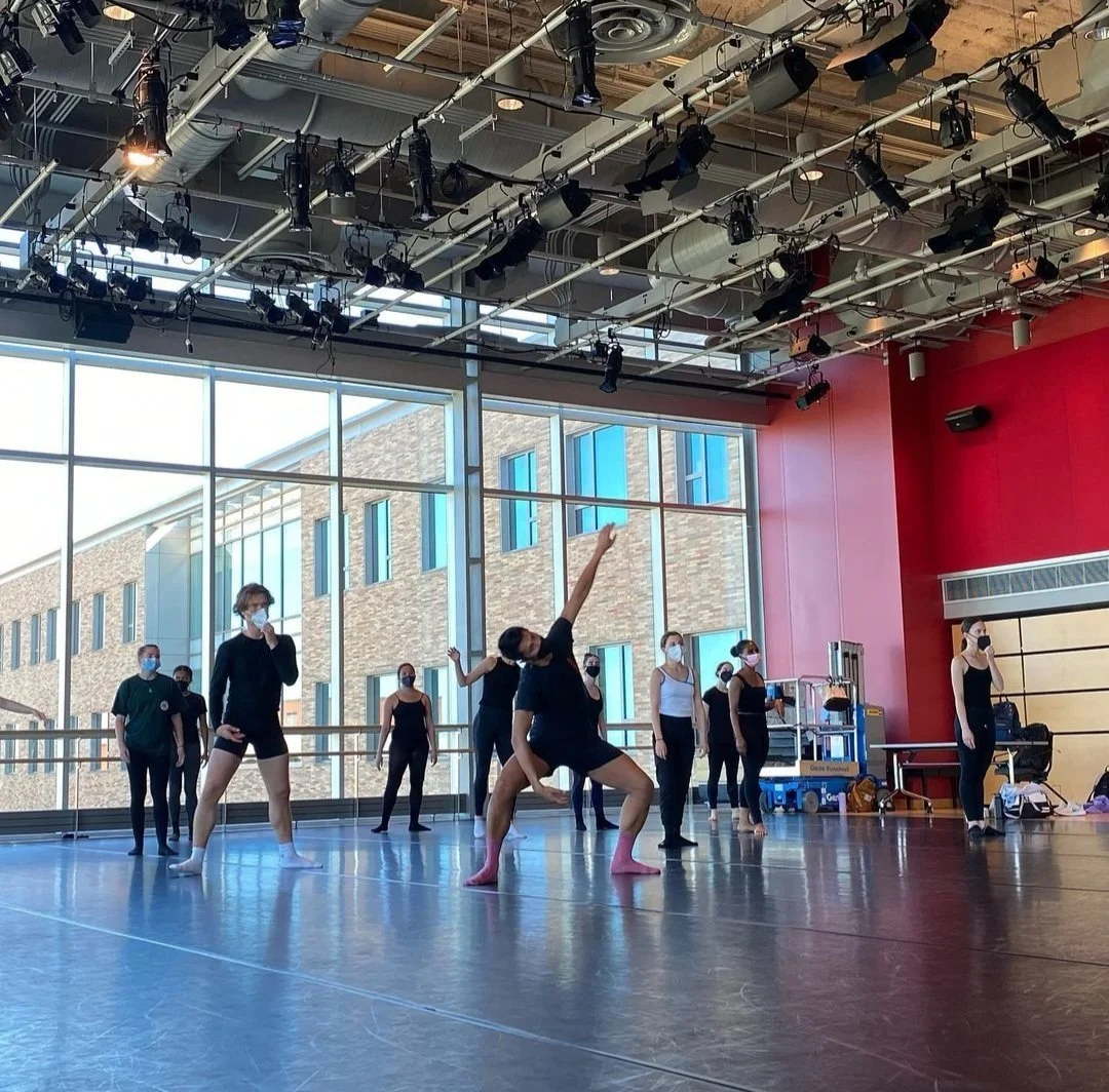 Dance rehearsal in a spacious studio with dancers wearing masks, large windows, and a red wall.