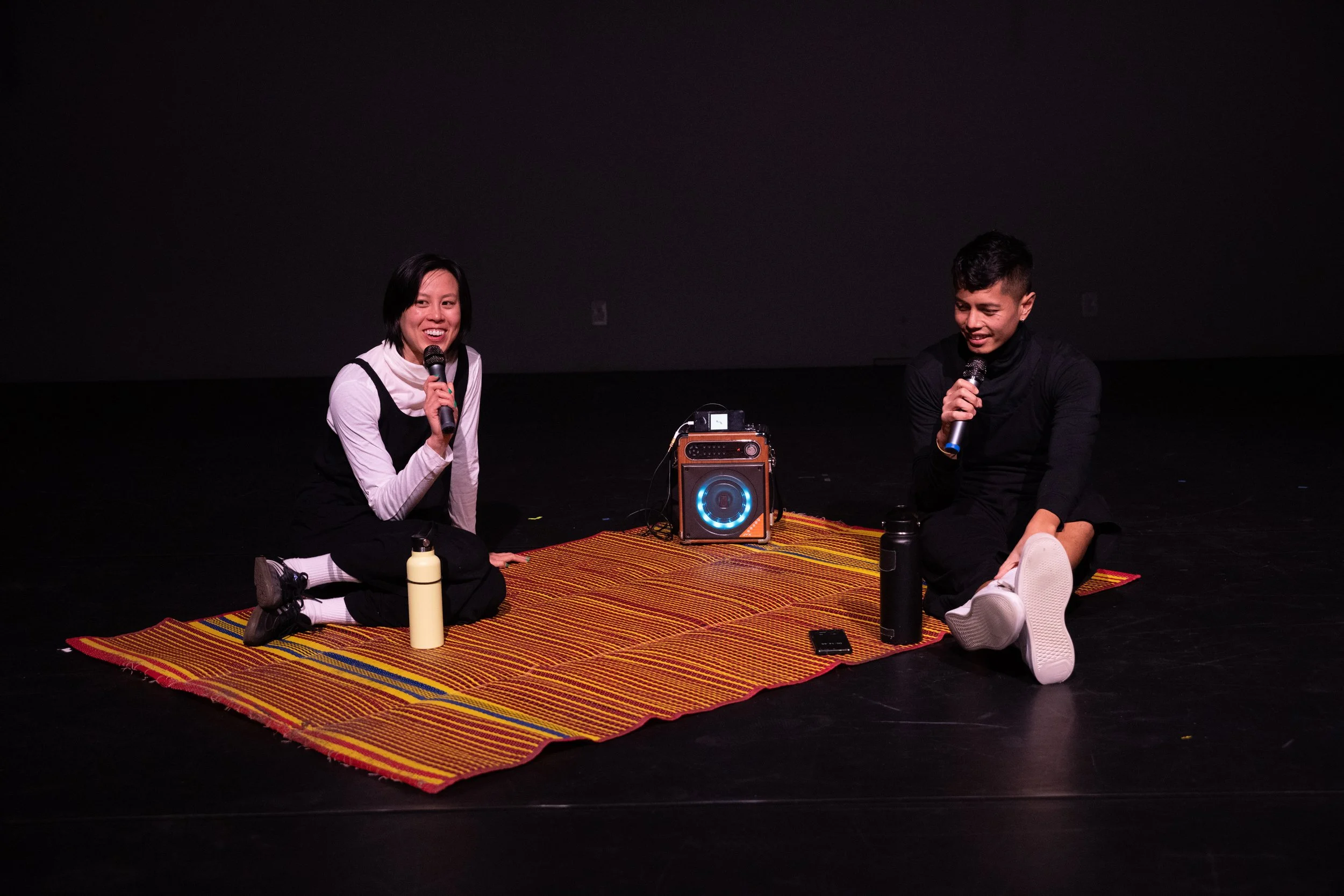 Two people sitting on a colorful rug, holding microphones, with a portable speaker and water bottles in front of them, in a dark stage setting.