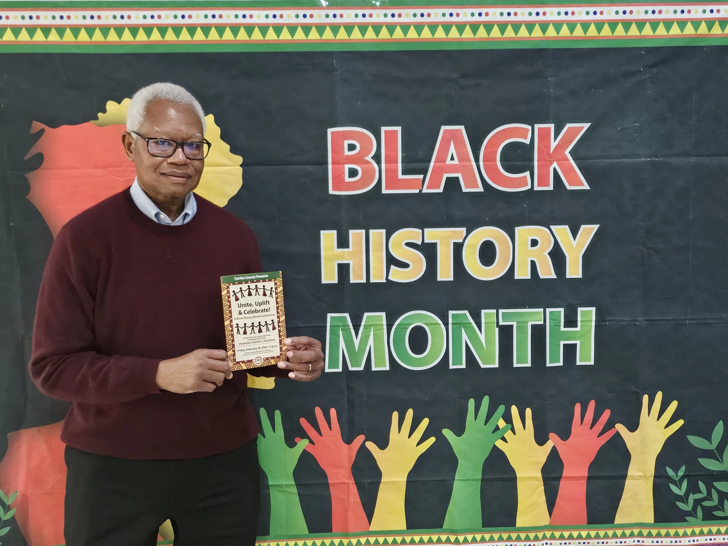 An elderly man with gray hair, wearing glasses, a maroon sweater, and a white collared shirt, standing in front of a colorful banner for Black History Month, holding a flyer that promotes unity, uplift, and celebration.