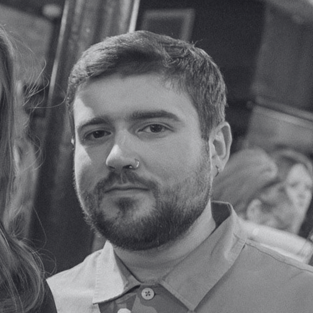 A young man with short hair and a beard, wearing a collared shirt with a nose piercing, looking at the camera in a black and white photo.