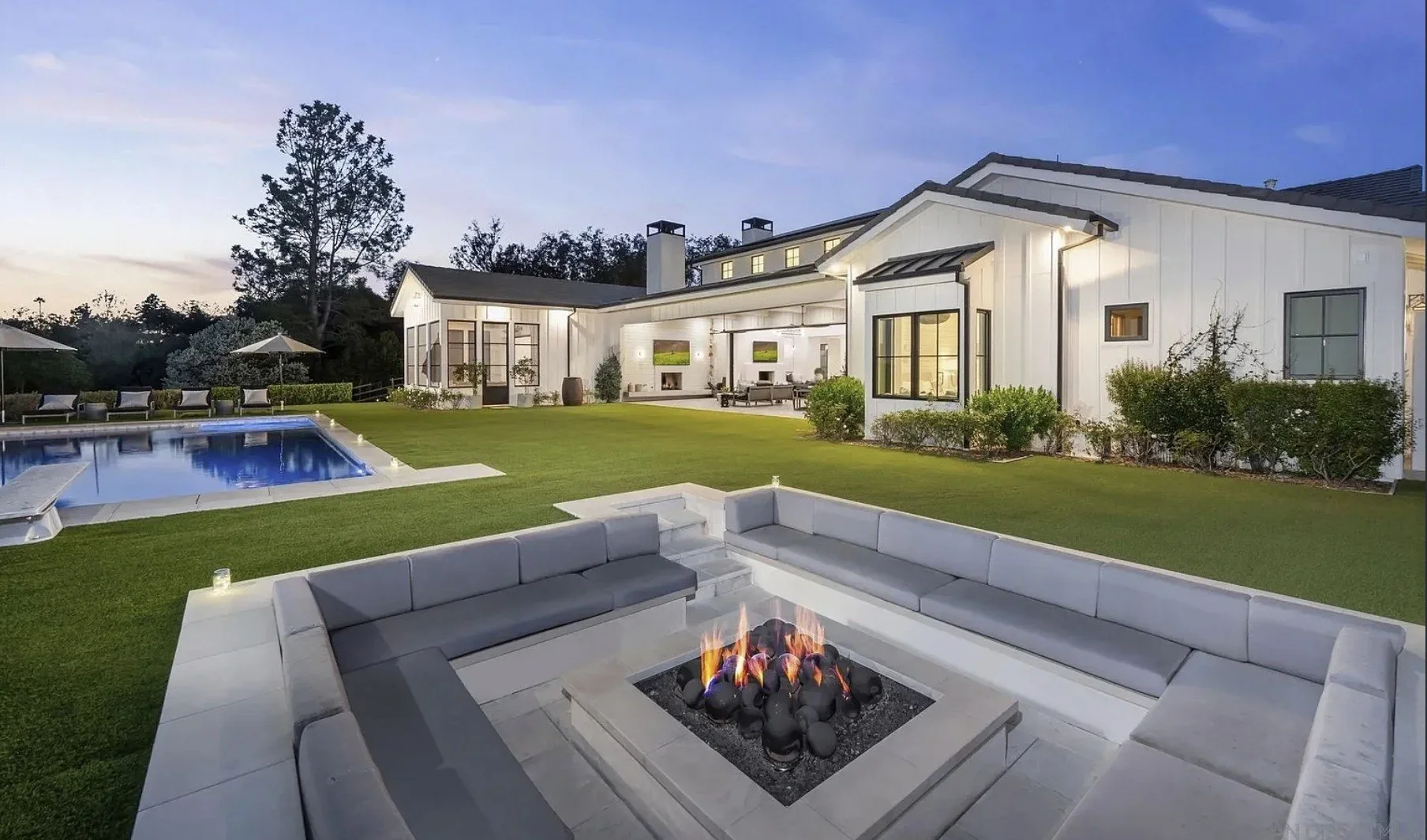 A modern backyard with a rectangular pool, outdoor seating around a fire pit, a well-manicured lawn, and a large white house with large windows and outdoor living area at dusk.