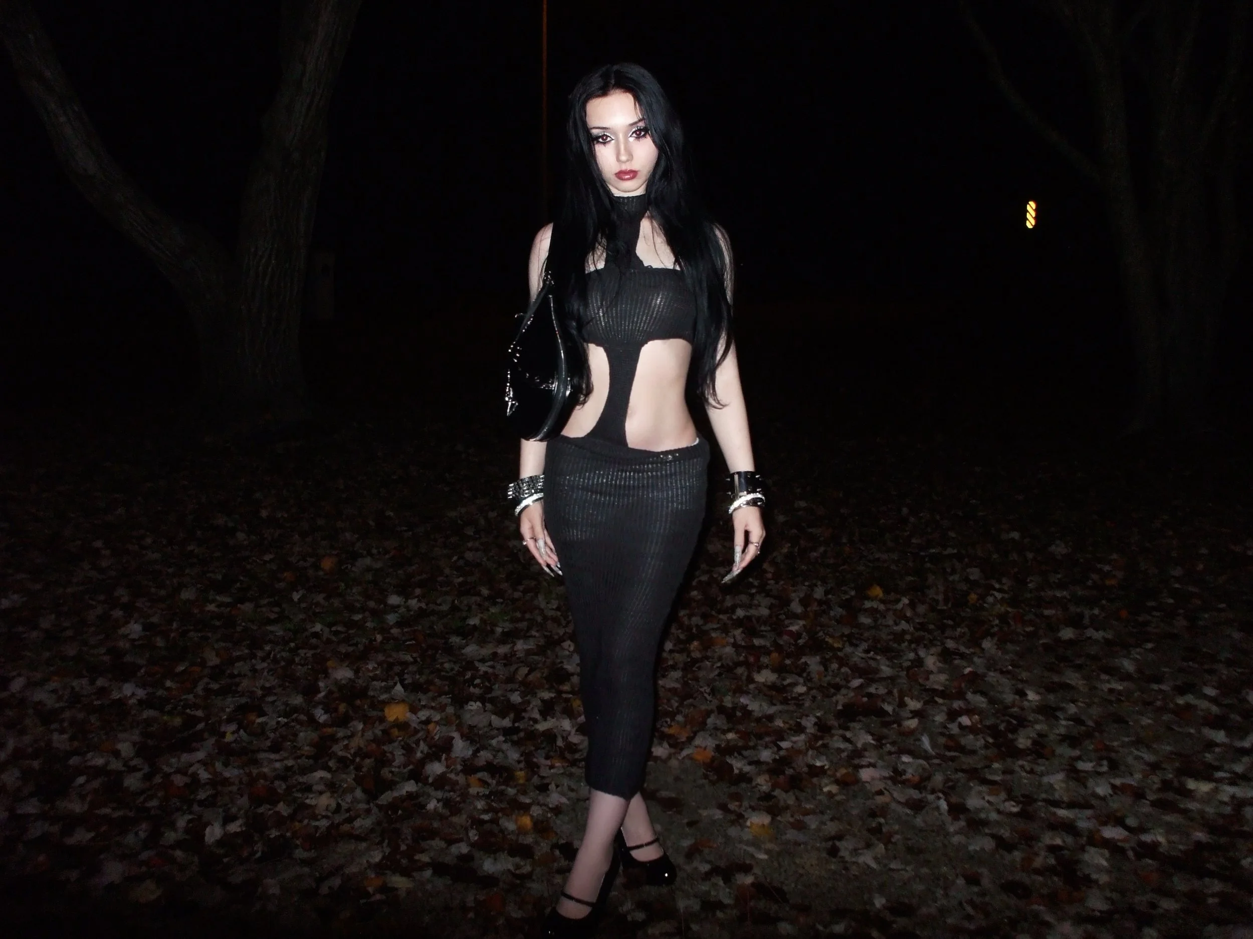 A woman with long black hair and dark makeup standing on a dark, wooded path at night, dressed in a black, custom black Megagatha one-of-one Cross Dress.