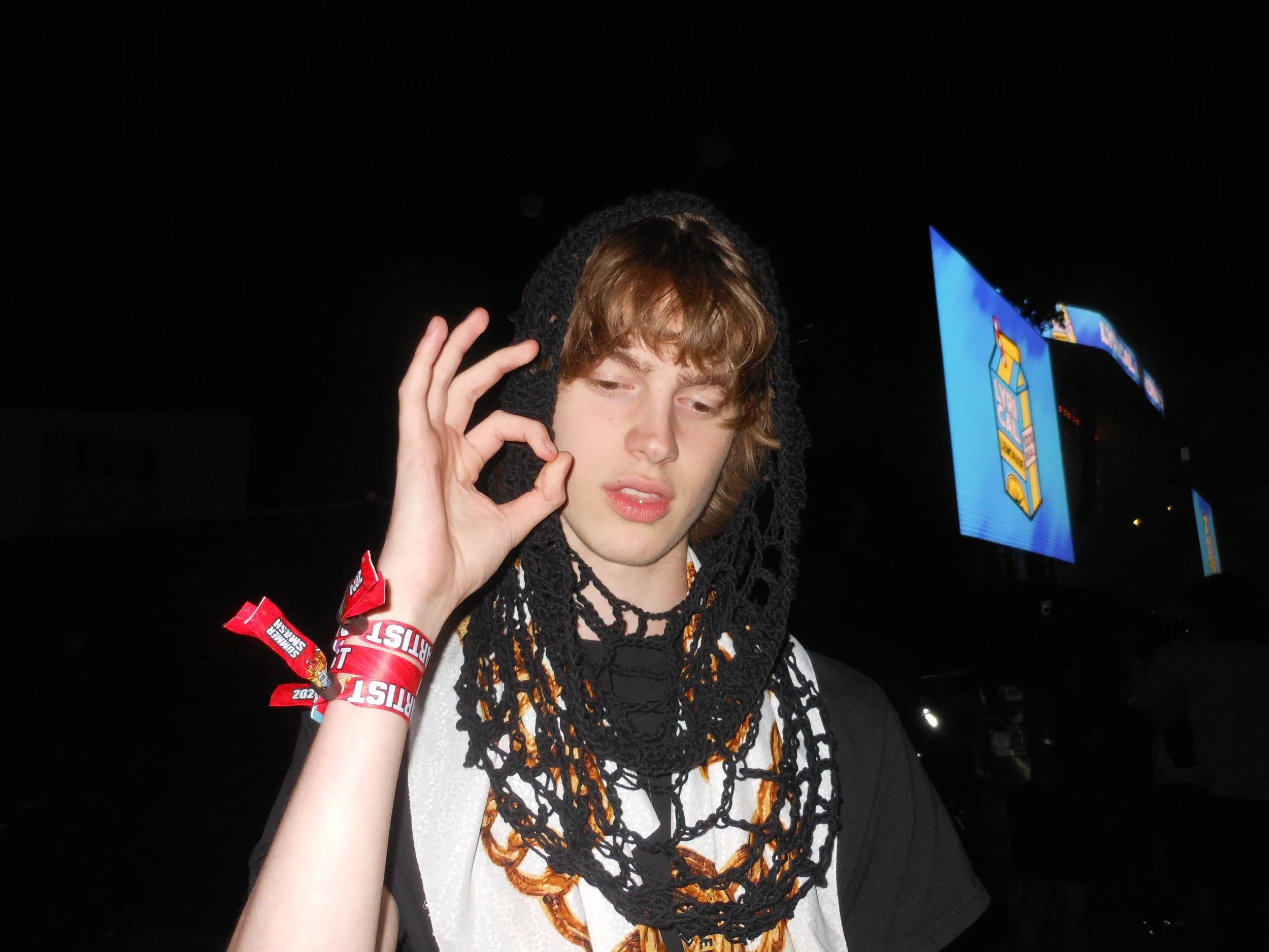 A young man with curly brown hair making an OK gesture with his right hand, wearing a Megagatha black crochet Spiderweb Hood and multiple black chain necklaces, in a dark setting with LED screens in the background at Summer Smash Festival in Chicago.