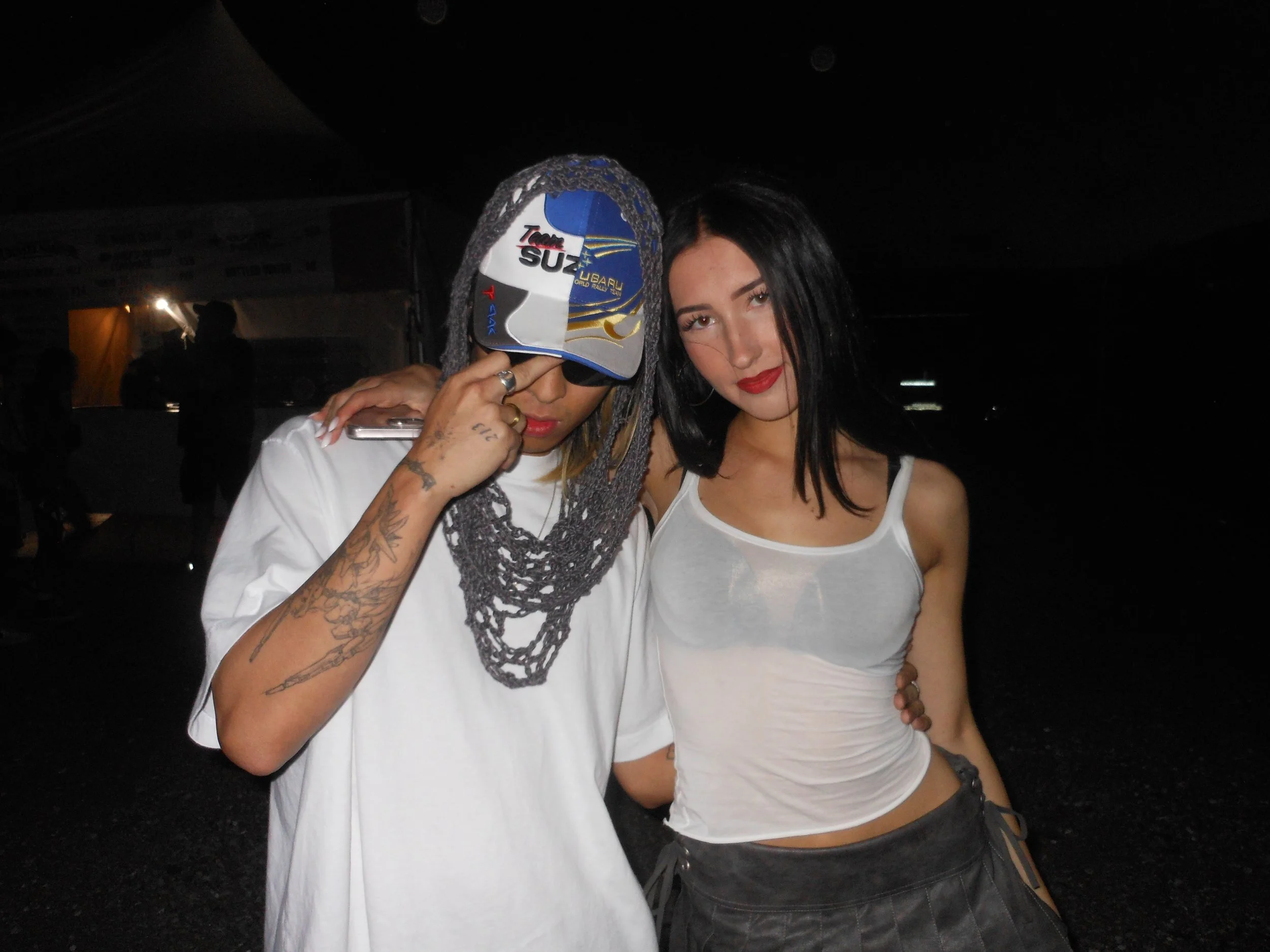 Two people posing together, one wearing a white t-shirt, sunglasses, a cap, and a Megagatha grey crochet Spiderweb Hood, the other wearing a white tank top and a black skirt, in a dark outdoor setting at Summer Smash Festival in Chicago.