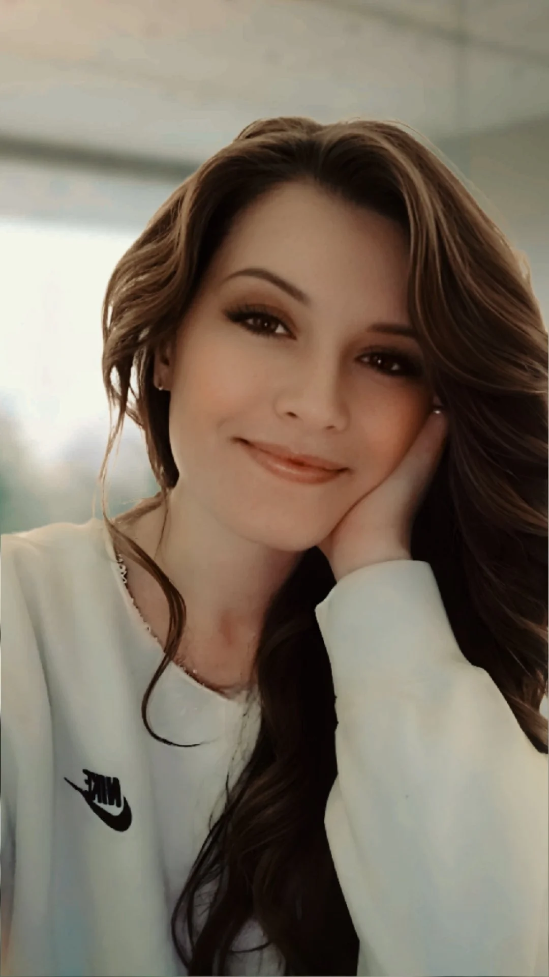 Close-up of a smiling woman with long brown hair, resting her face on her hand, wearing a white Nike sweatshirt.