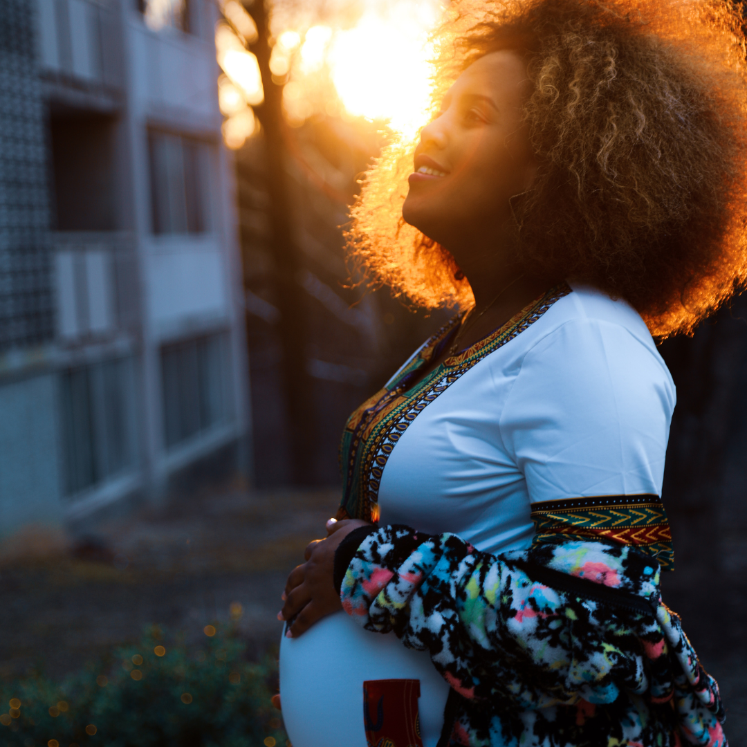 A pregnant woman with curly hair smiling outdoors at sunset, wearing a white dress with colorful embroidery and a multicolored fleece jacket, holding her belly.