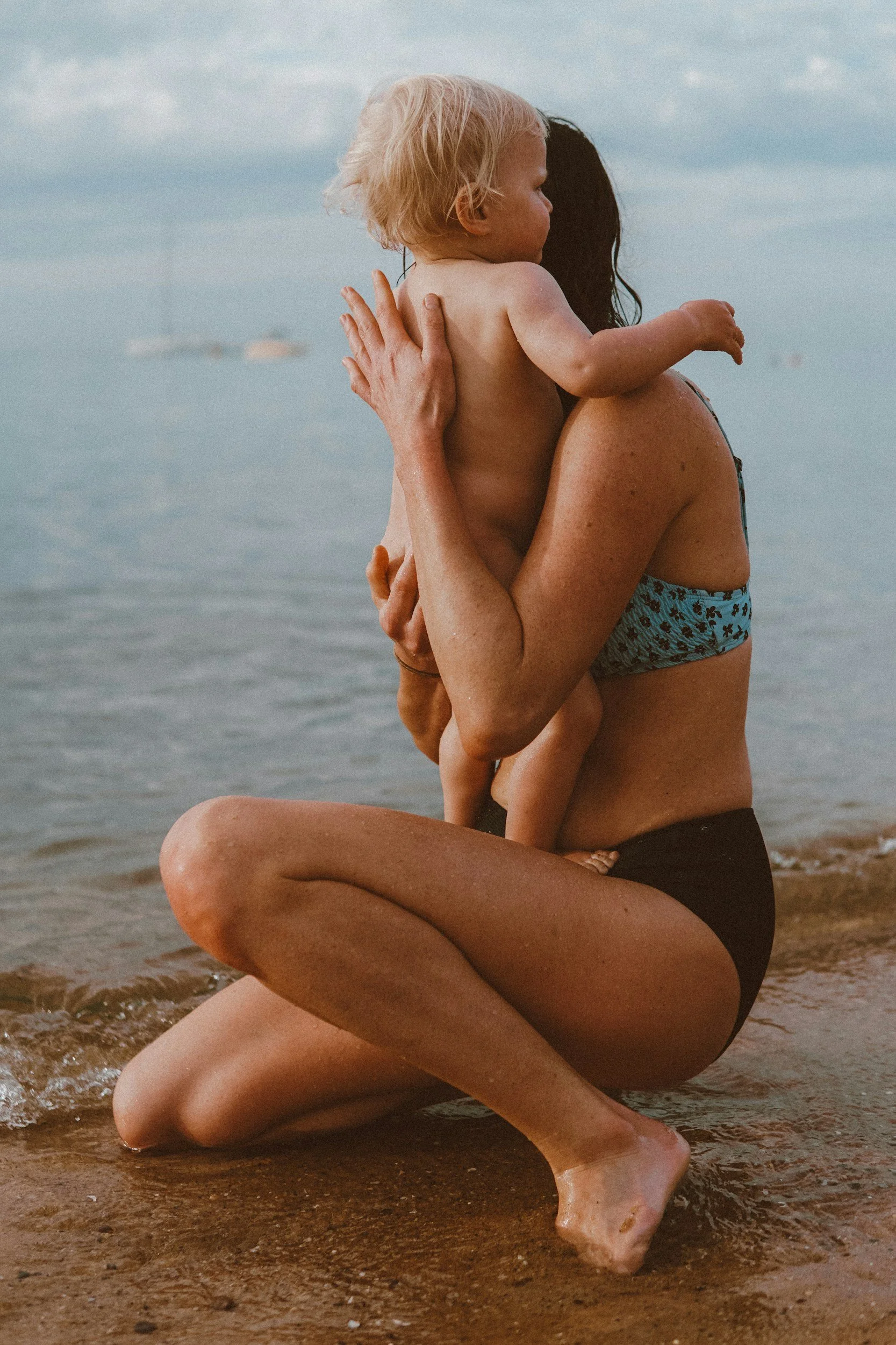 A woman kneeling on the beach while holding and hugging a small child, both with wet skin, near the shoreline with water and cloudy sky in the background.
