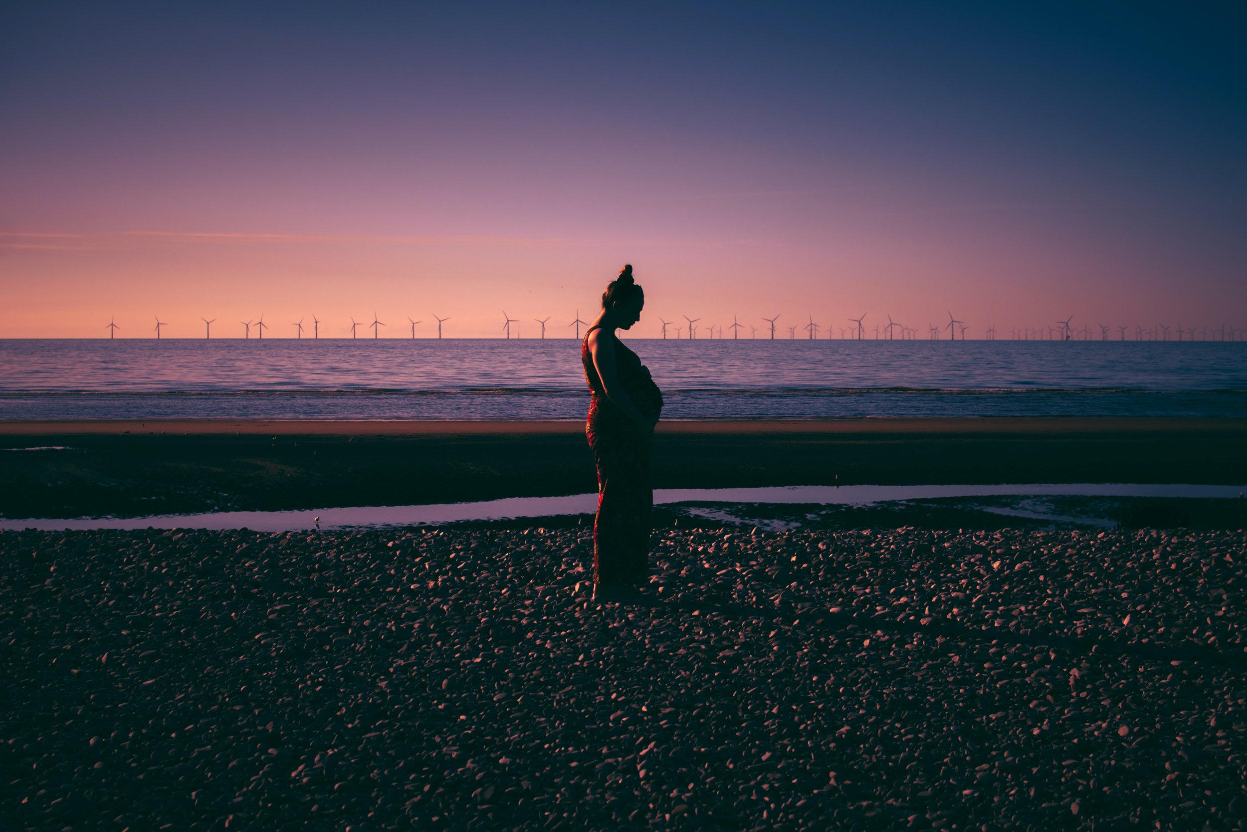 Silhouette of a pregnant woman standing on a beach during sunset, with wind turbines visible in the distance.