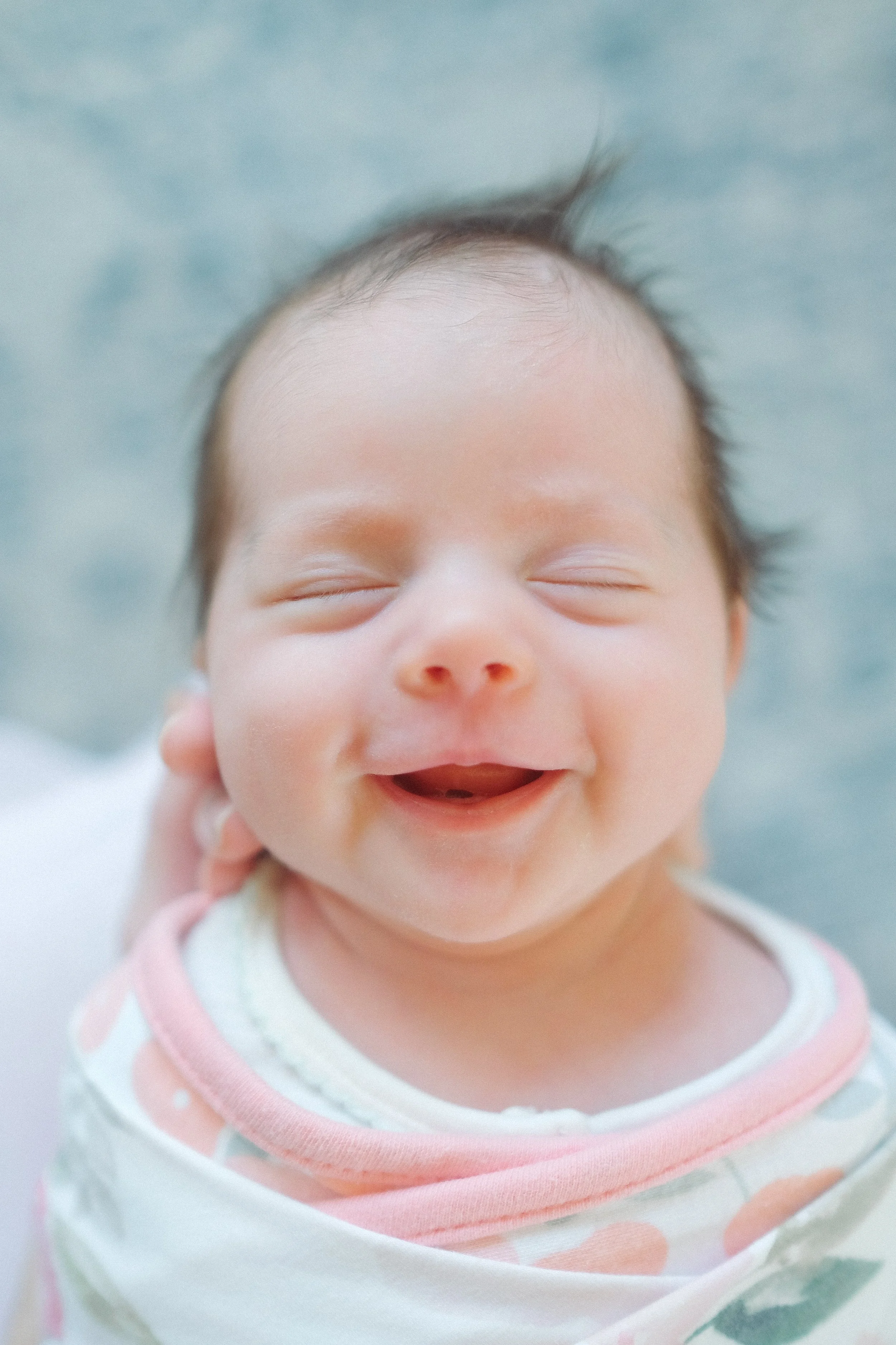 A close-up image of a smiling baby with closed eyes, wearing a white and pink patterned shirt, on a soft background.
