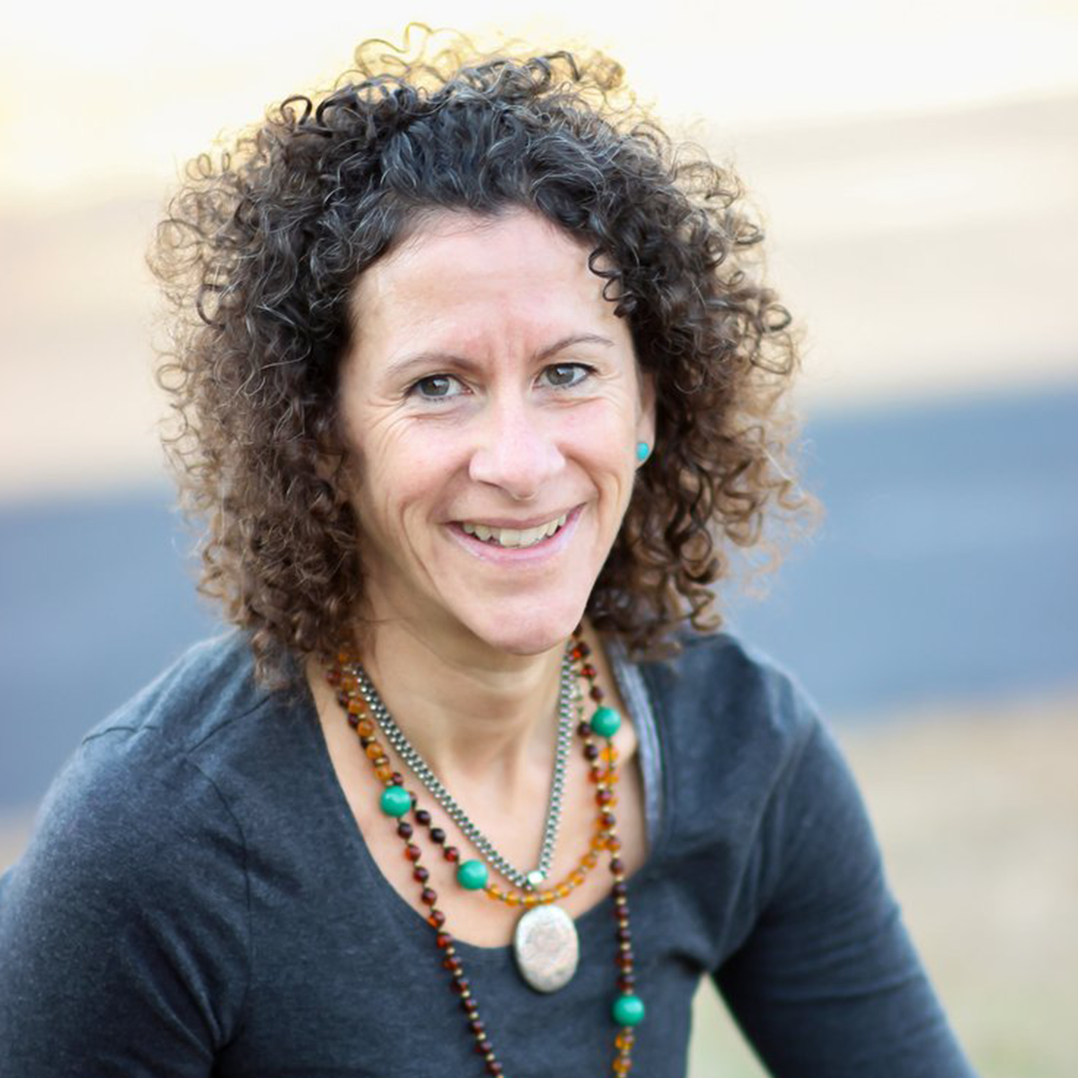 A woman (Rebecca Goodwin) with curly brown hair smiling outdoors, wearing a black top and layered necklaces with turquoise beads and a large pendant.
