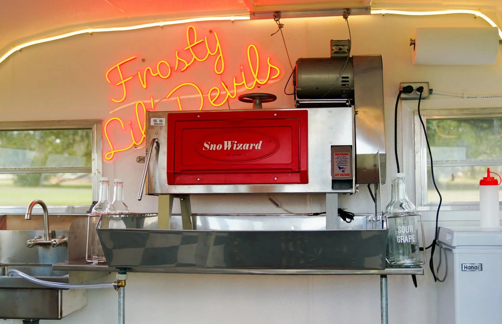A food truck kitchen with a neon sign. Inside, there is a red Sno Wizard ice shaver machine and several glass bottles. There is also a condiment squeeze bottle on a counter to the side.