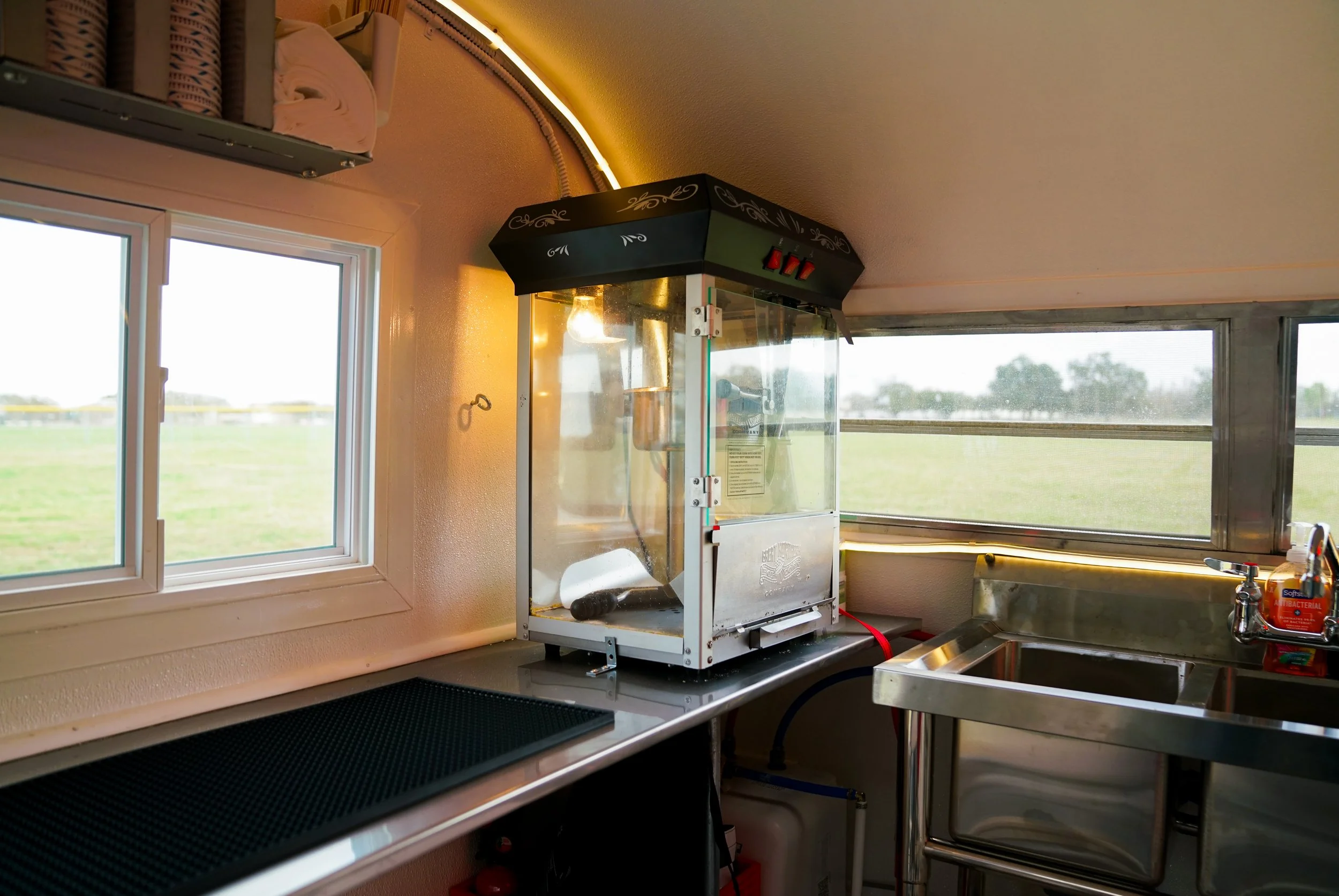 A popcorn machine on a countertop next to a window in a small kitchen or concession stand, with a stainless steel sink and soap dispenser nearby.