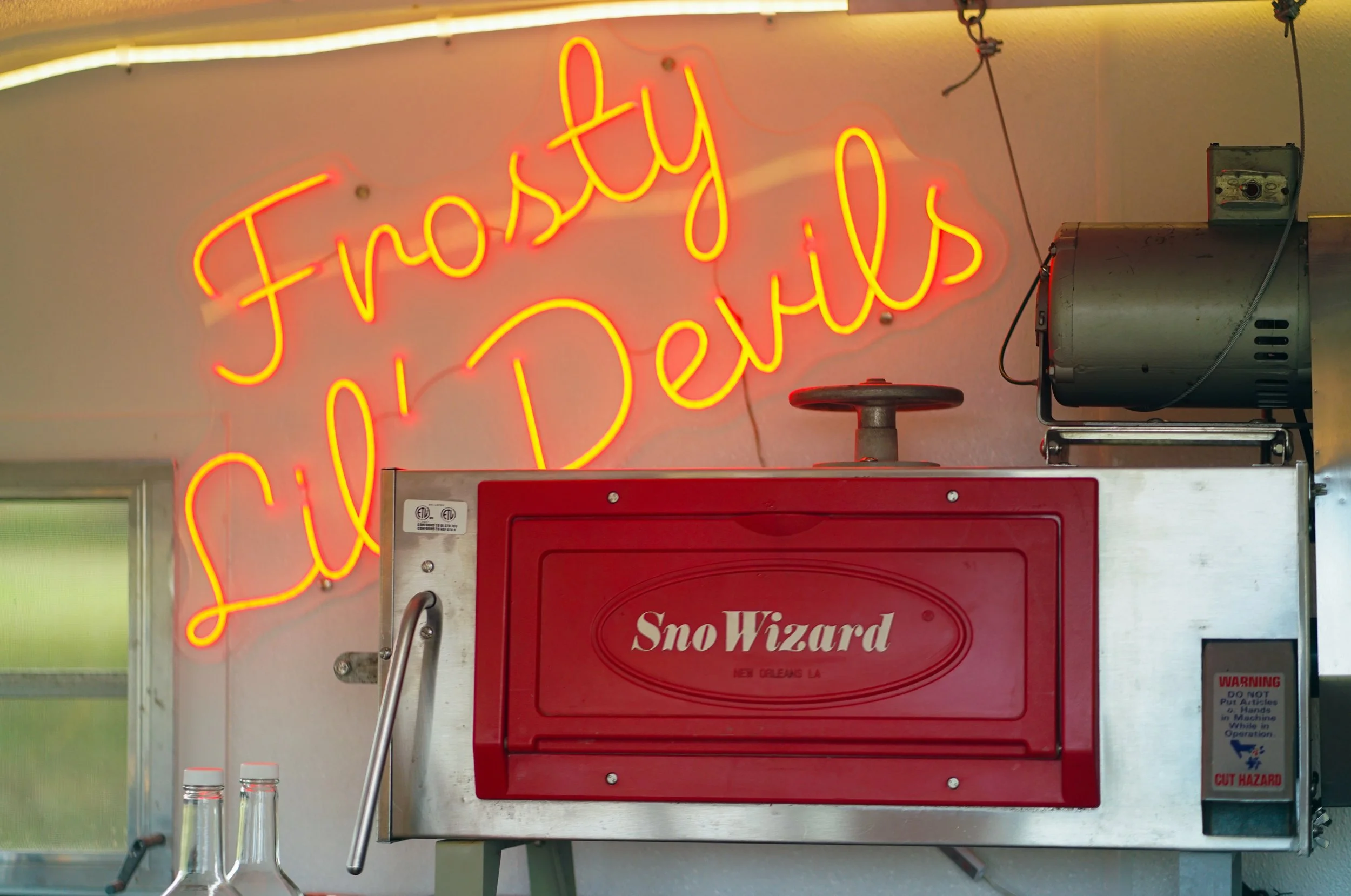 Neon sign in yellow and red reading 'Frosty Lil Devils' hangs above a red SnoWizard ice shaver machine in a food stand.