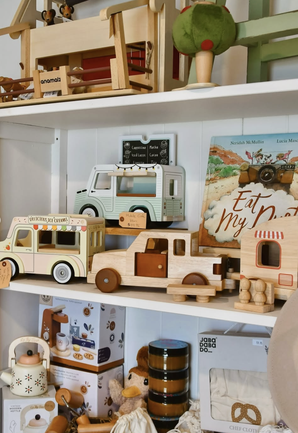 Display of wooden toy vehicles including a vintage ice cream truck and a delivery truck, along with other decorative items like books, plush toys, and small accessories on store shelves.