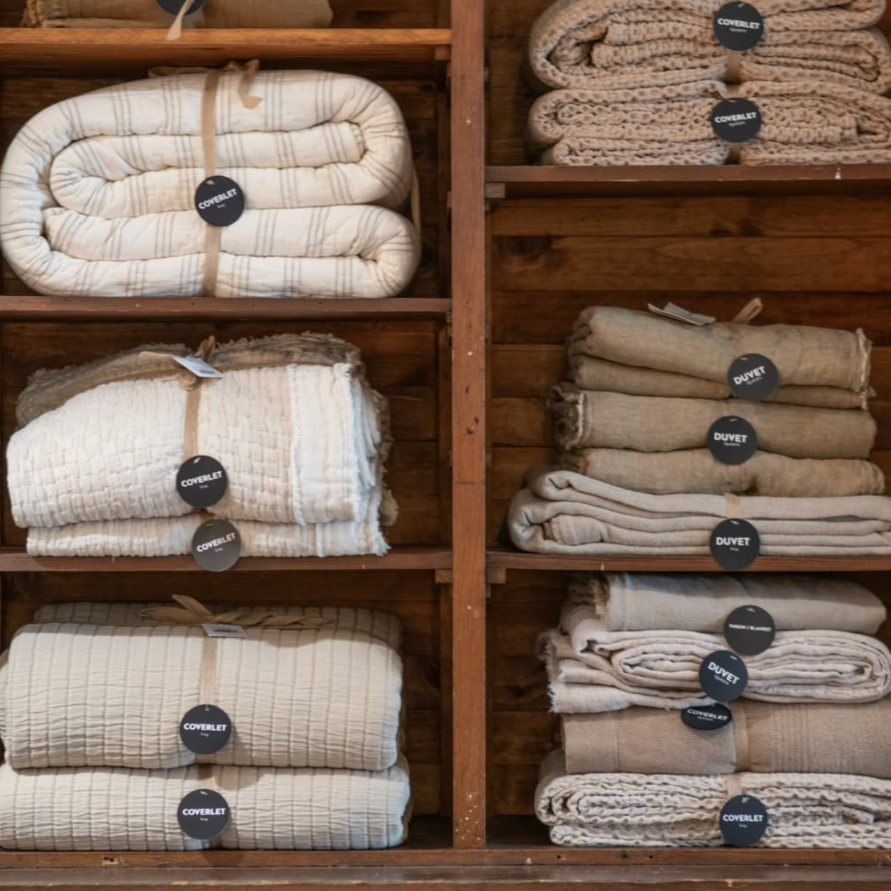 Stacks of beige and cream-colored blankets and throws on wooden shelves, labeled with tags reading 'coverlet' and 'duvet'.
