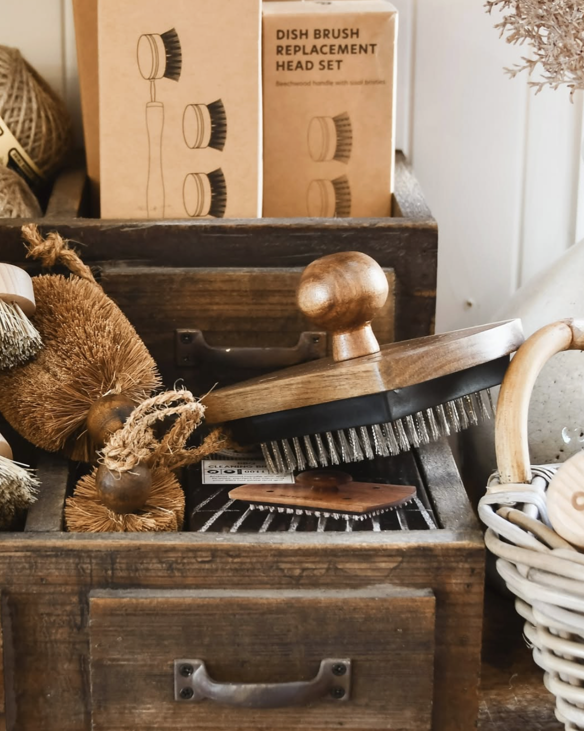 A wooden drawer filled with cleaning brushes, including natural bristle brushes, a brush with metal bristles, and a wooden-handled brush, with a box of dish brush replacement heads in the background.