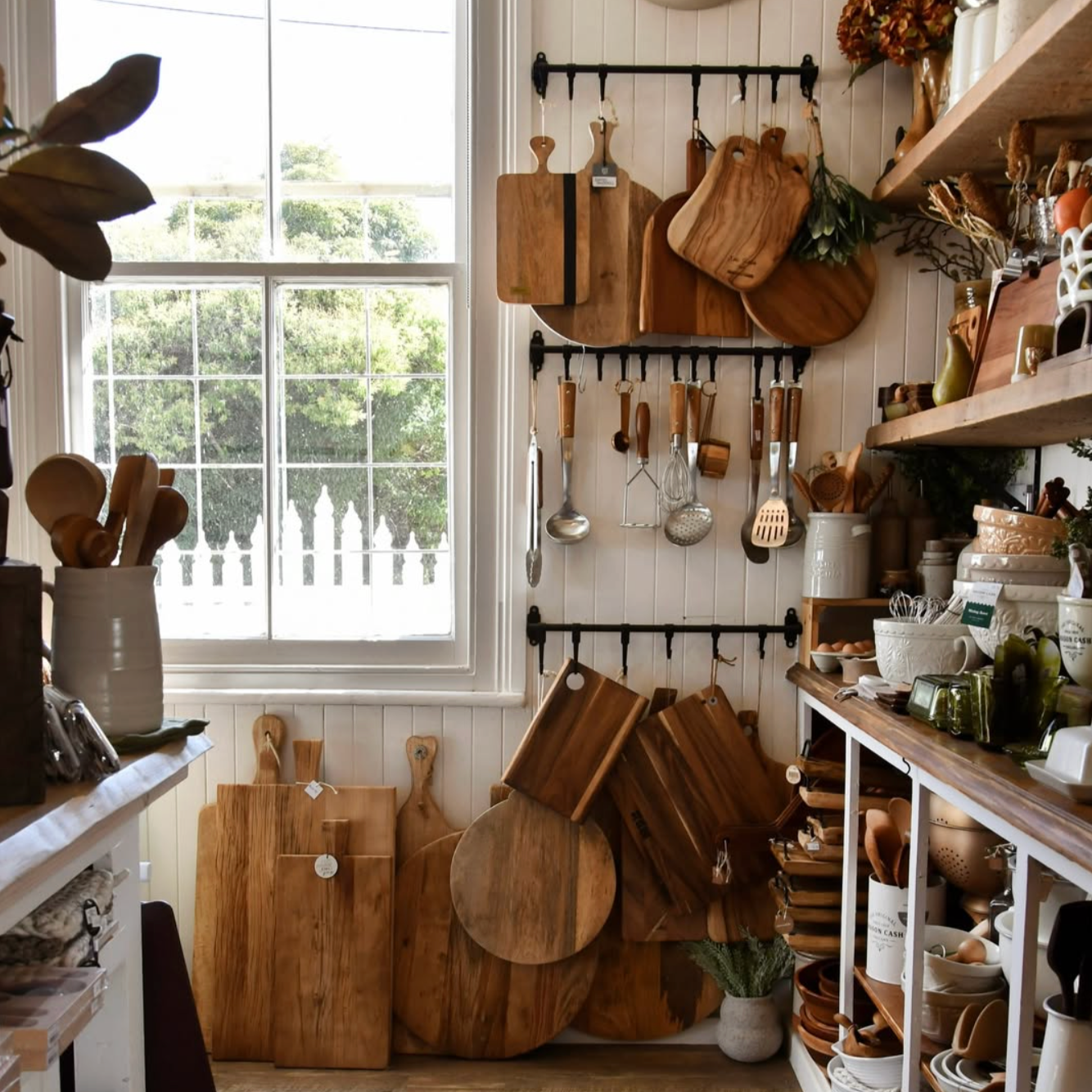 A cozy kitchen corner with wooden cutting boards, utensils, and pottery on shelves and wall hooks, illuminated by natural light from a large window.
