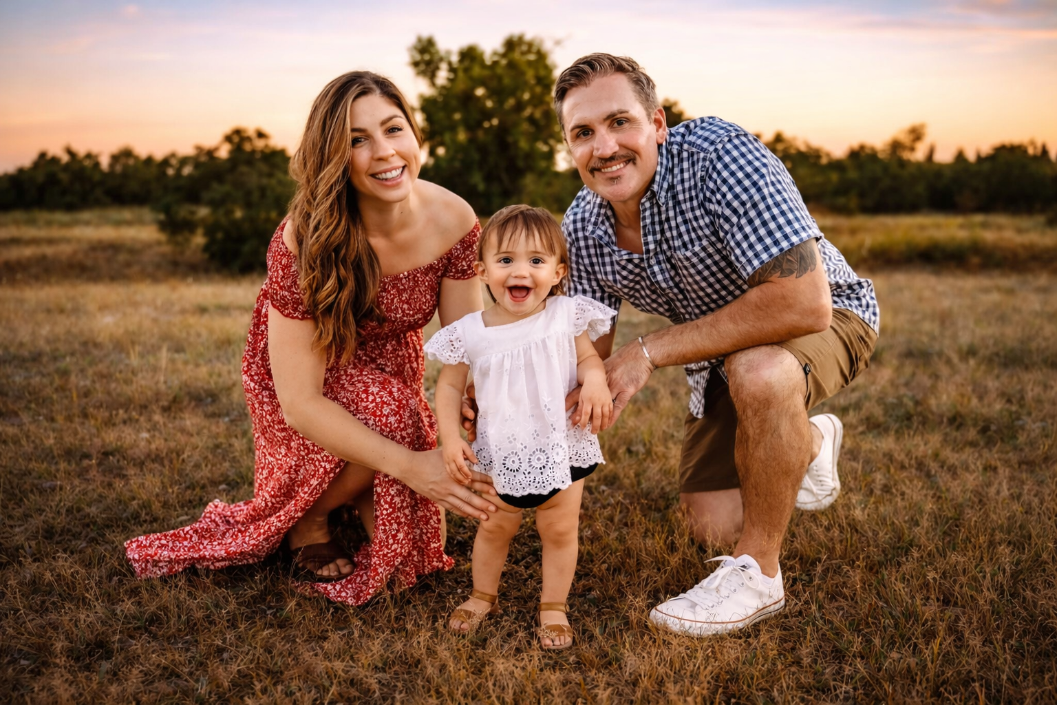 A smiling family of three, a mother, father, and young daughter, outdoors on a grassy field during sunset, all crouching and facing the camera.