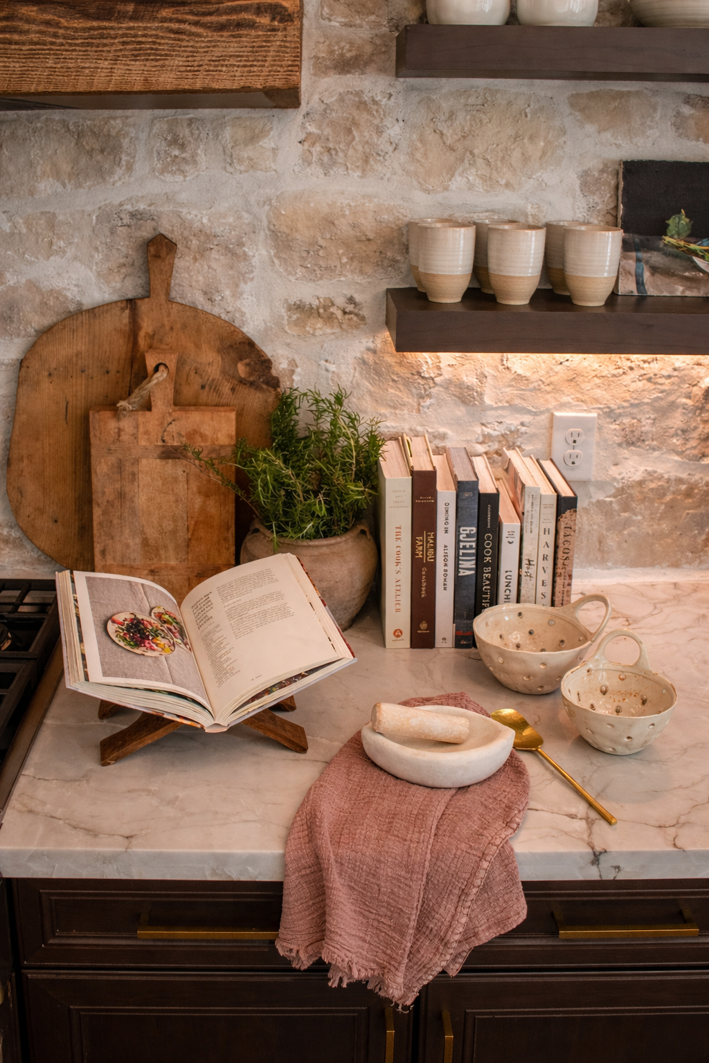 A kitchen countertop with a cookbook on a wooden stand, a potted plant, stacked books, a pink cloth, a marble mortar and pestle, two ceramic bowls, a gold spoon, and a marble tray. There are floating shelves with white cups and bowls, and a stone wall in the background.