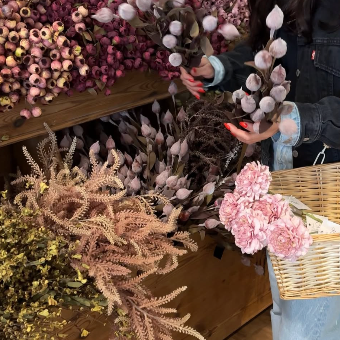Person holding a bundle of pink and brown dried flowers, surrounded by other pink, peach, and purple floral arrangements on display.