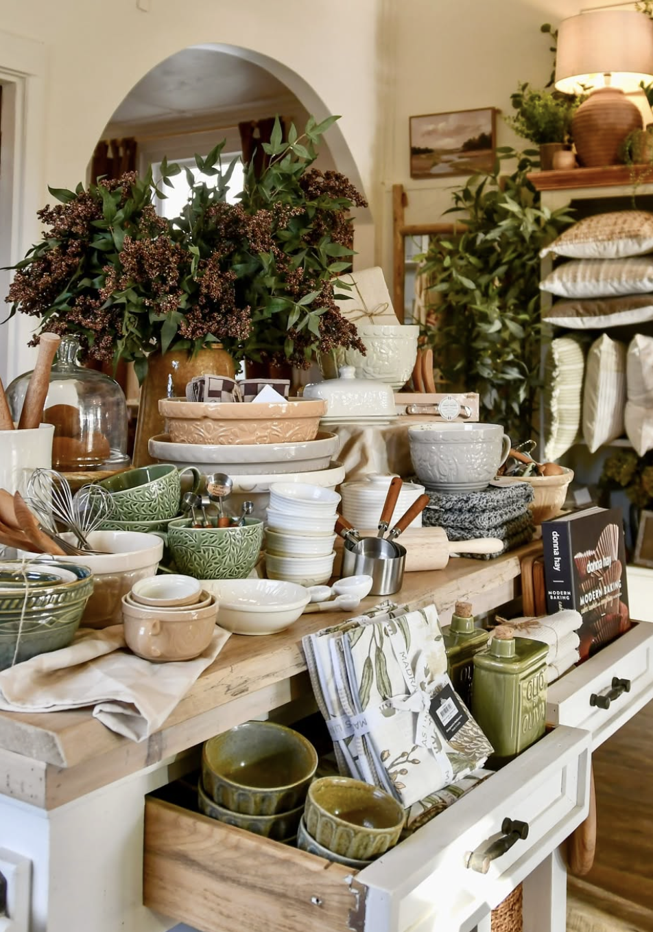 Display of various kitchenware including bowls, cups, and utensils on a wooden countertop surrounded by decorative plants and home decor items.