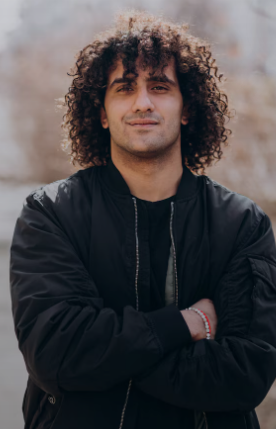 A man with curly hair wearing a black jacket and standing outdoors with arms crossed.