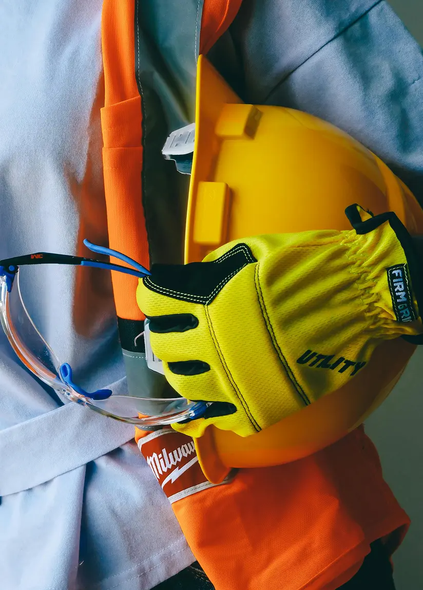 Close-up of personal safety gear including yellow gloves marked 'UTIMA', clear safety glasses with black and blue frames, a yellow helmet, and a high-visibility orange jacket with a white logo, on a gray surface.