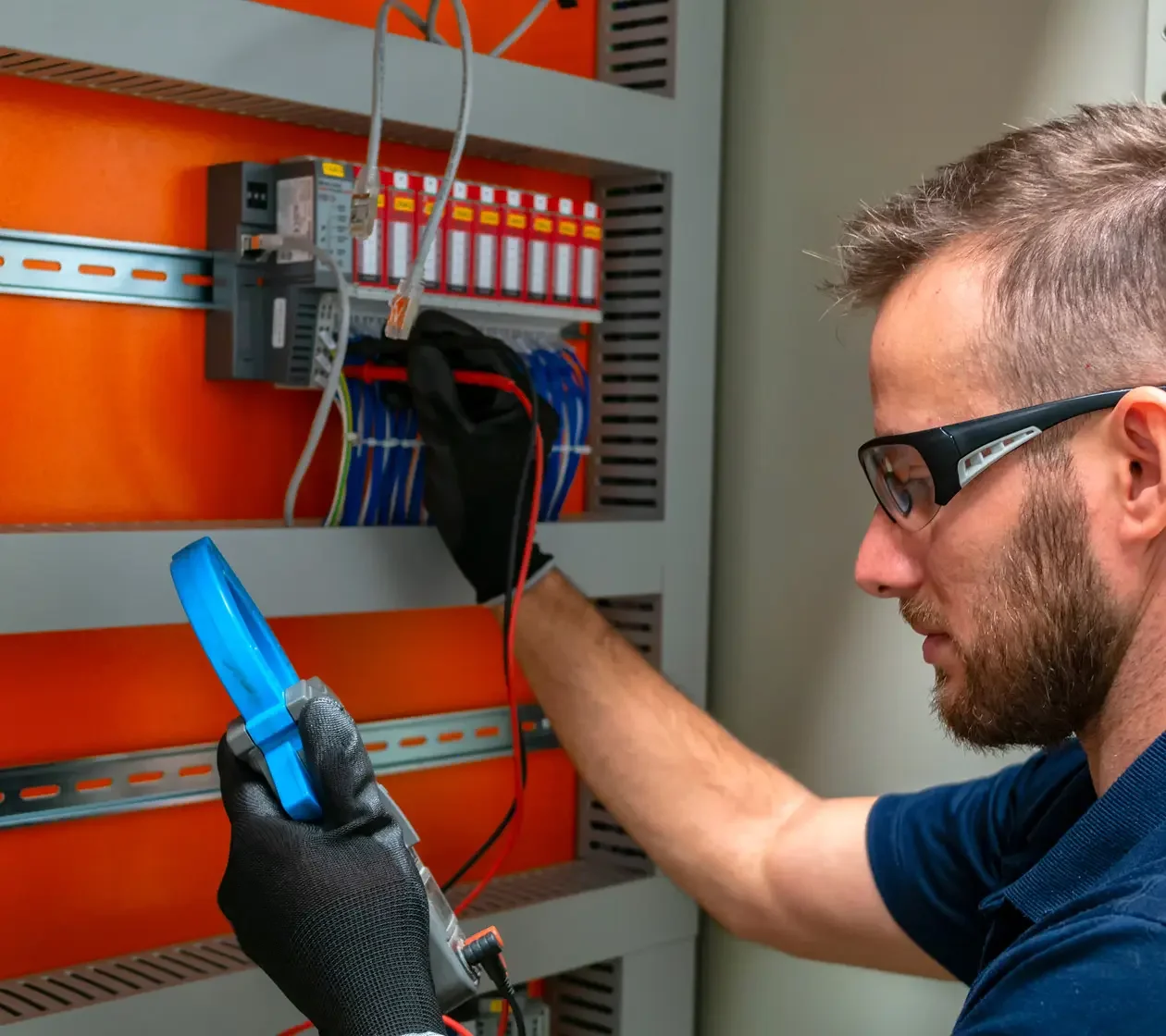 A man with glasses and a beard wearing a dark shirt, working on an electrical panel, holding a multimeter with a blue display, testing or troubleshooting electrical components.