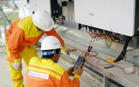Two electrical workers in safety gear inspecting solar inverter panels.