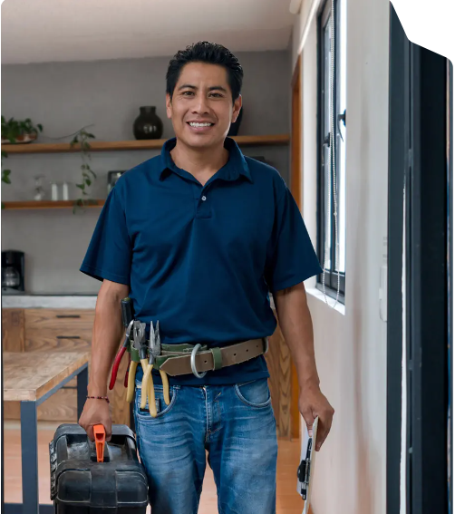 Man dressed in a blue polo shirt and jeans, carrying a toolbox and tools, standing inside a house near a window.