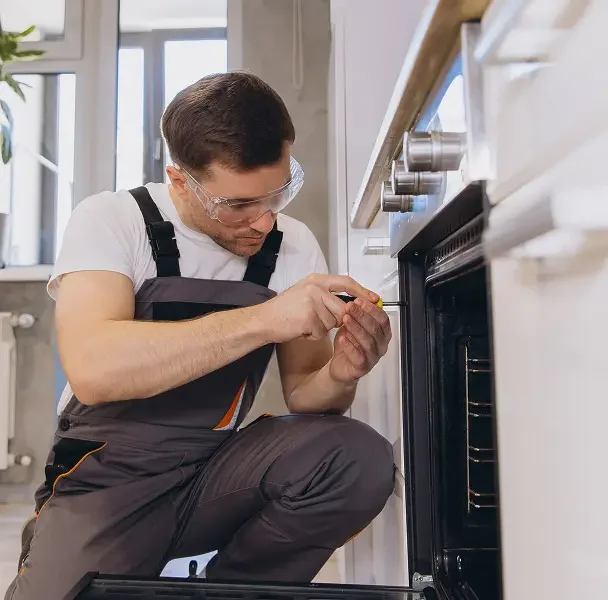A man wearing safety glasses, white shirt, and gray work overalls kneels on the floor and examines or repairs an oven in a kitchen.