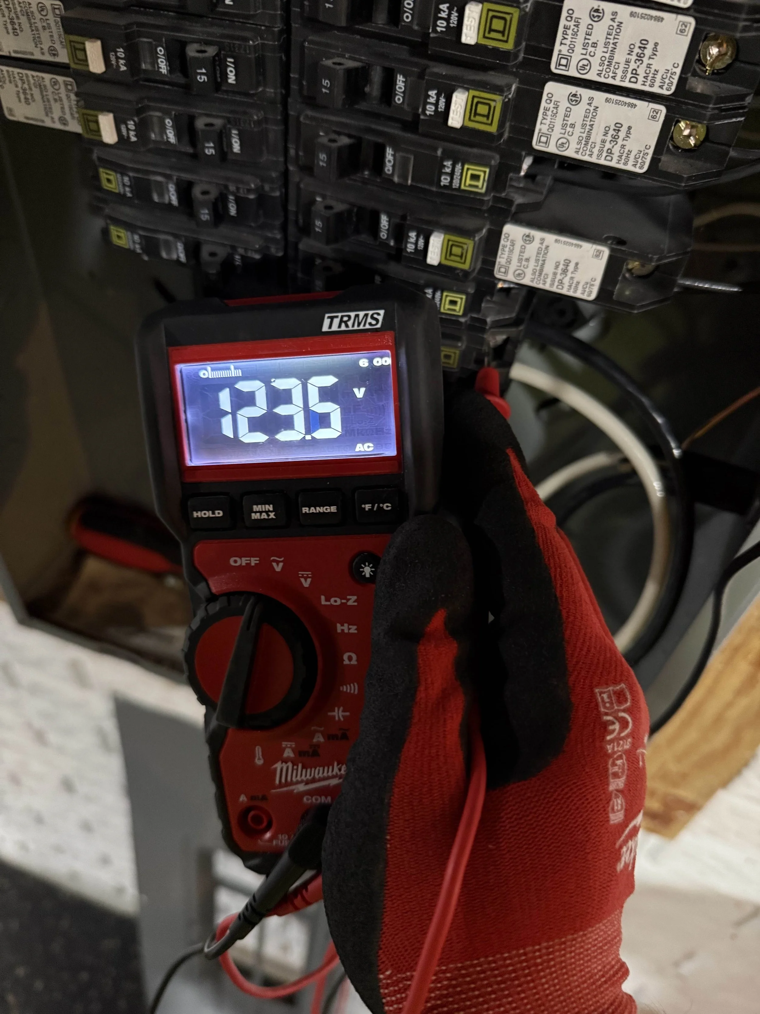 A person wearing red and black gloves is using a digital multimeter to measure voltage in an electrical panel containing circuit breakers. The multimeter displays a reading of 123.6 volts.