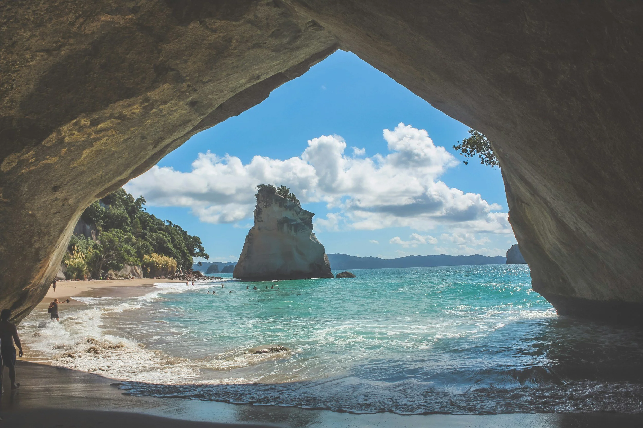 A view of a beach with turquoise water and a large limestone rock formation, seen through a natural rock archway with a cloudy sky and distant mountains.