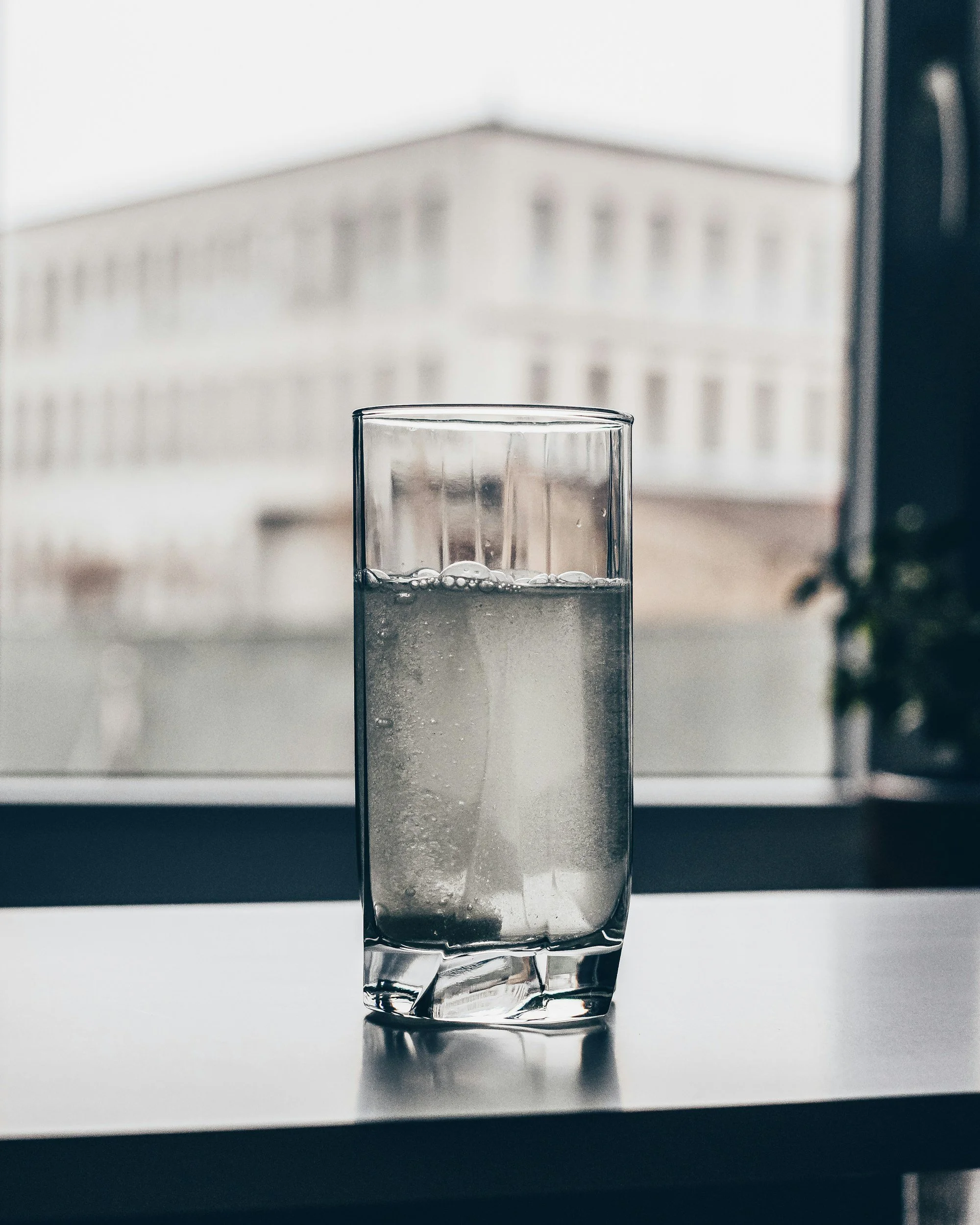 A clear glass of water with bubbles, placed on a windowsill or flat surface, with a distant building visible through the window in the background.