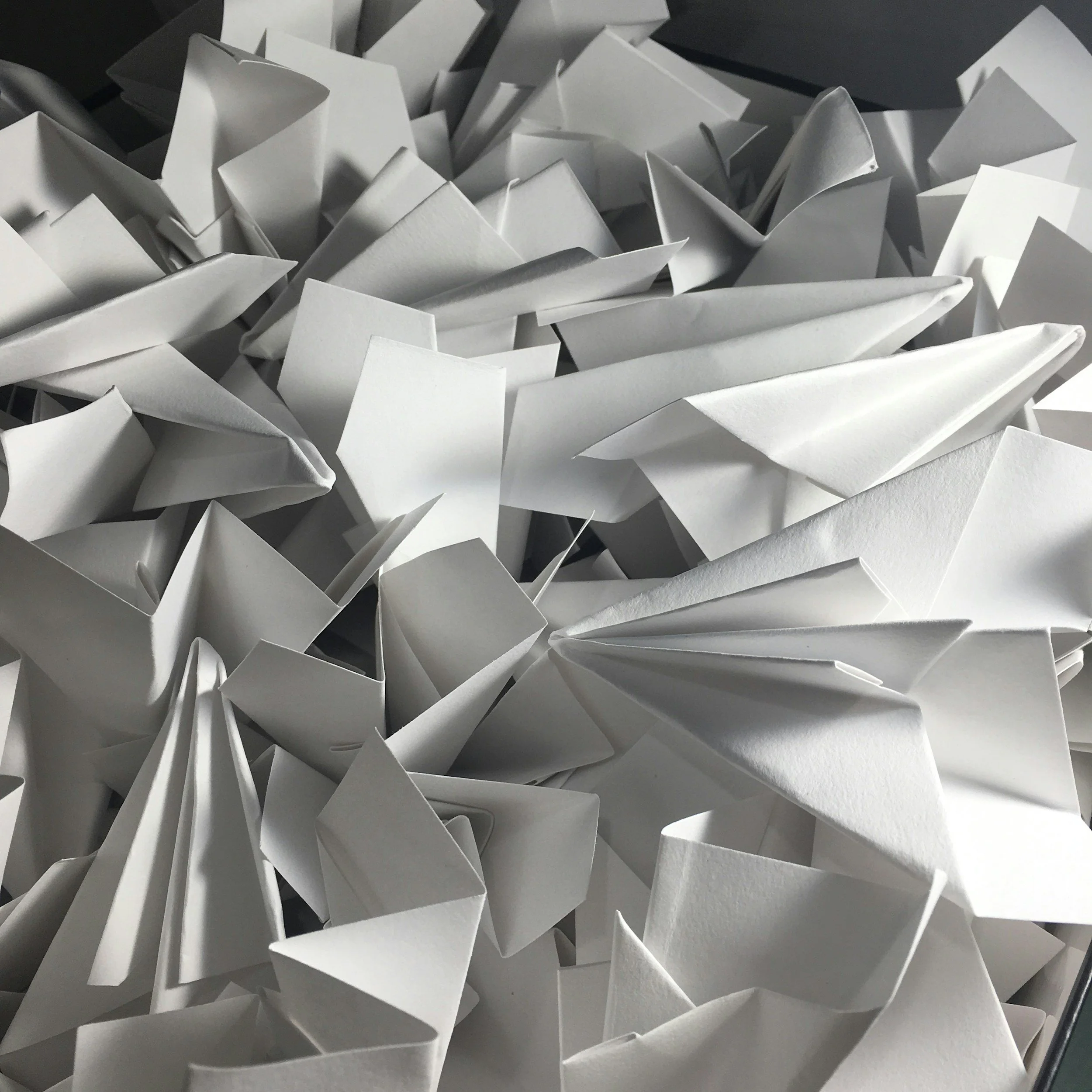 A close-up view of numerous white, folded paper boats made from paper of slightly different sizes and shapes, piled together.