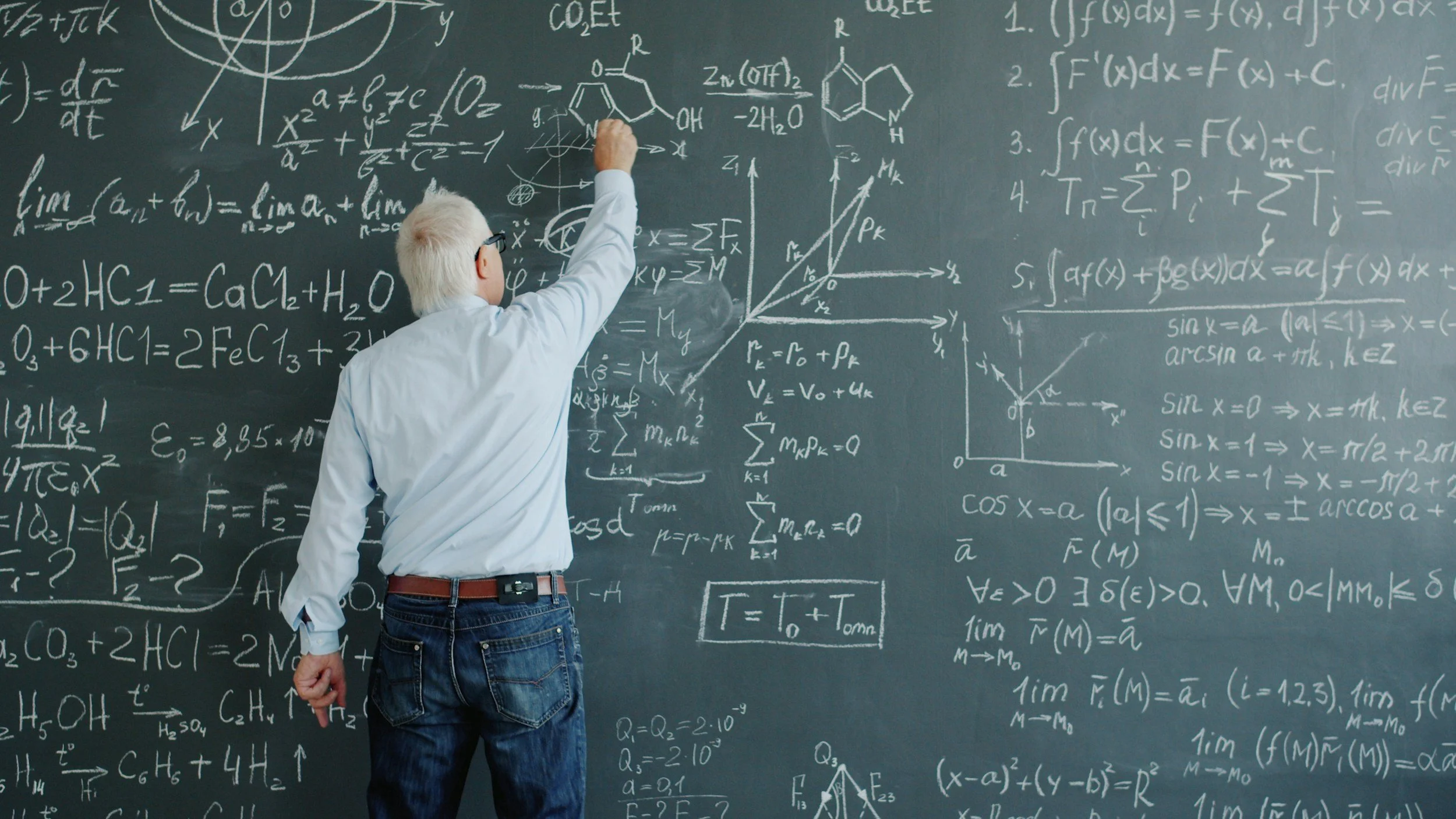 An elderly man with white hair and glasses, wearing a white shirt and jeans, writing complex equations and diagrams related to mathematics and science on a large chalkboard filled with chalk writing and sketches.