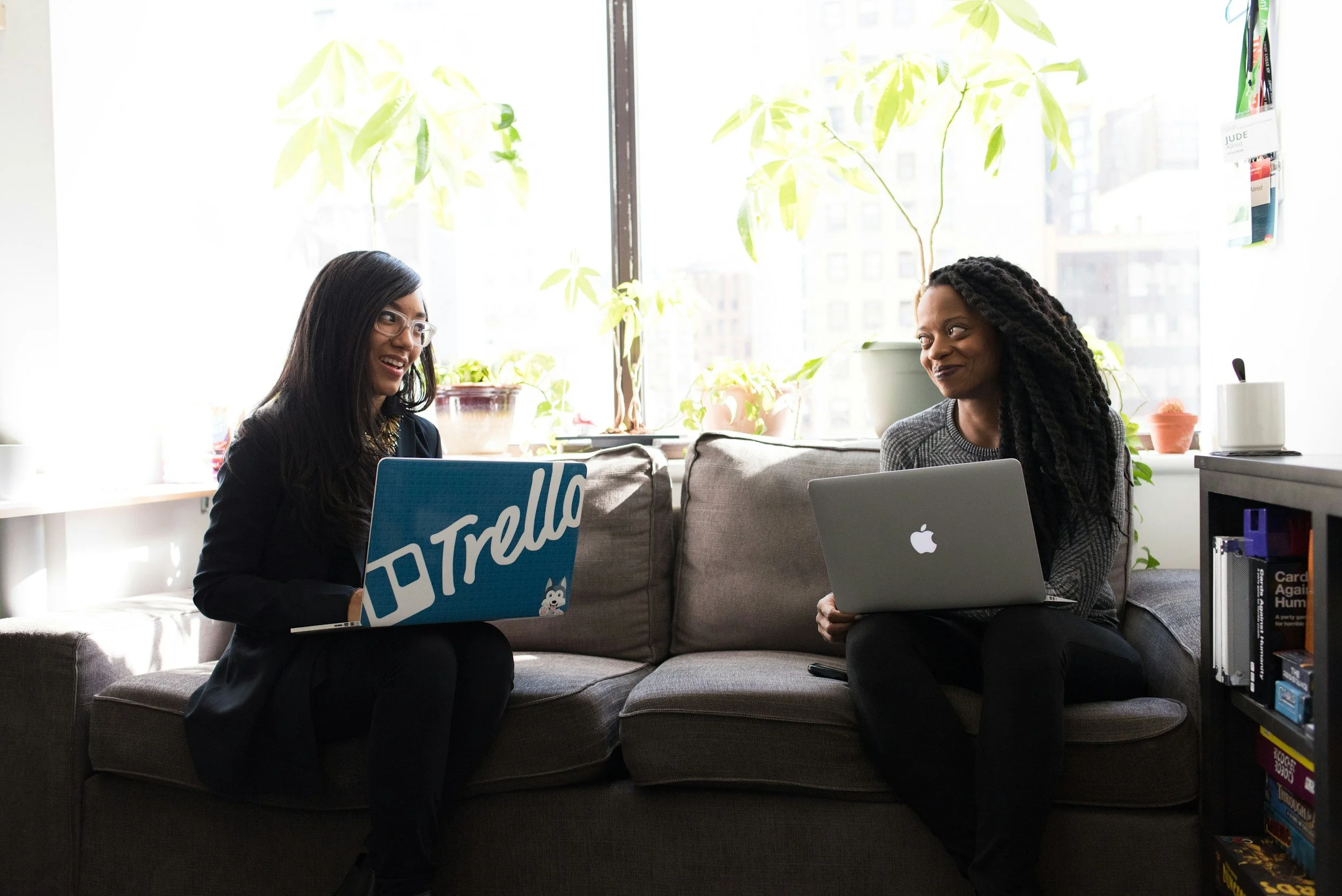 Two women sitting on a couch in an office, using laptops and engaging in conversation, with indoor plants and books around.